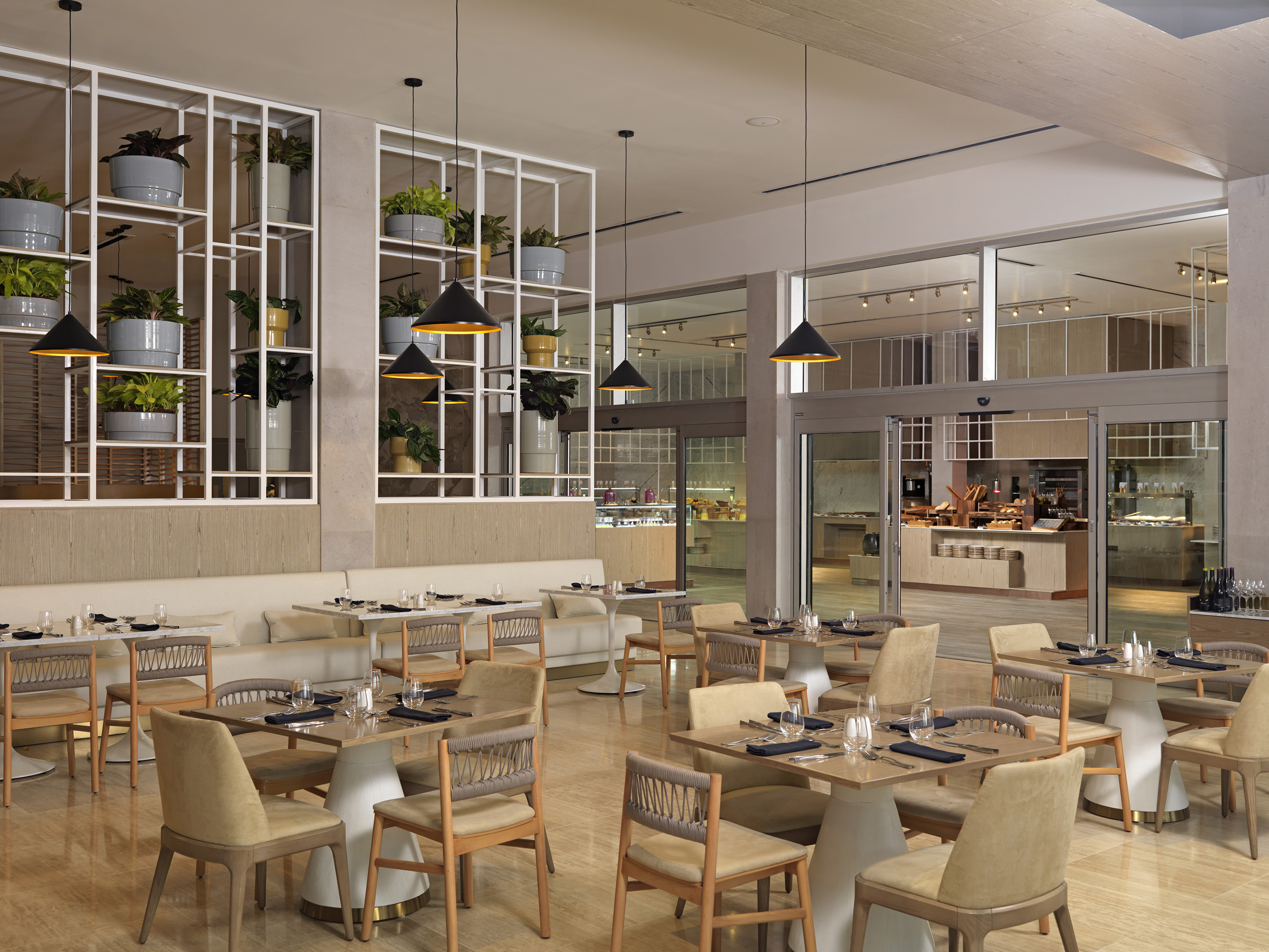 Modern restaurant dining area with set tables, tan chairs, pendant lights, and plants.