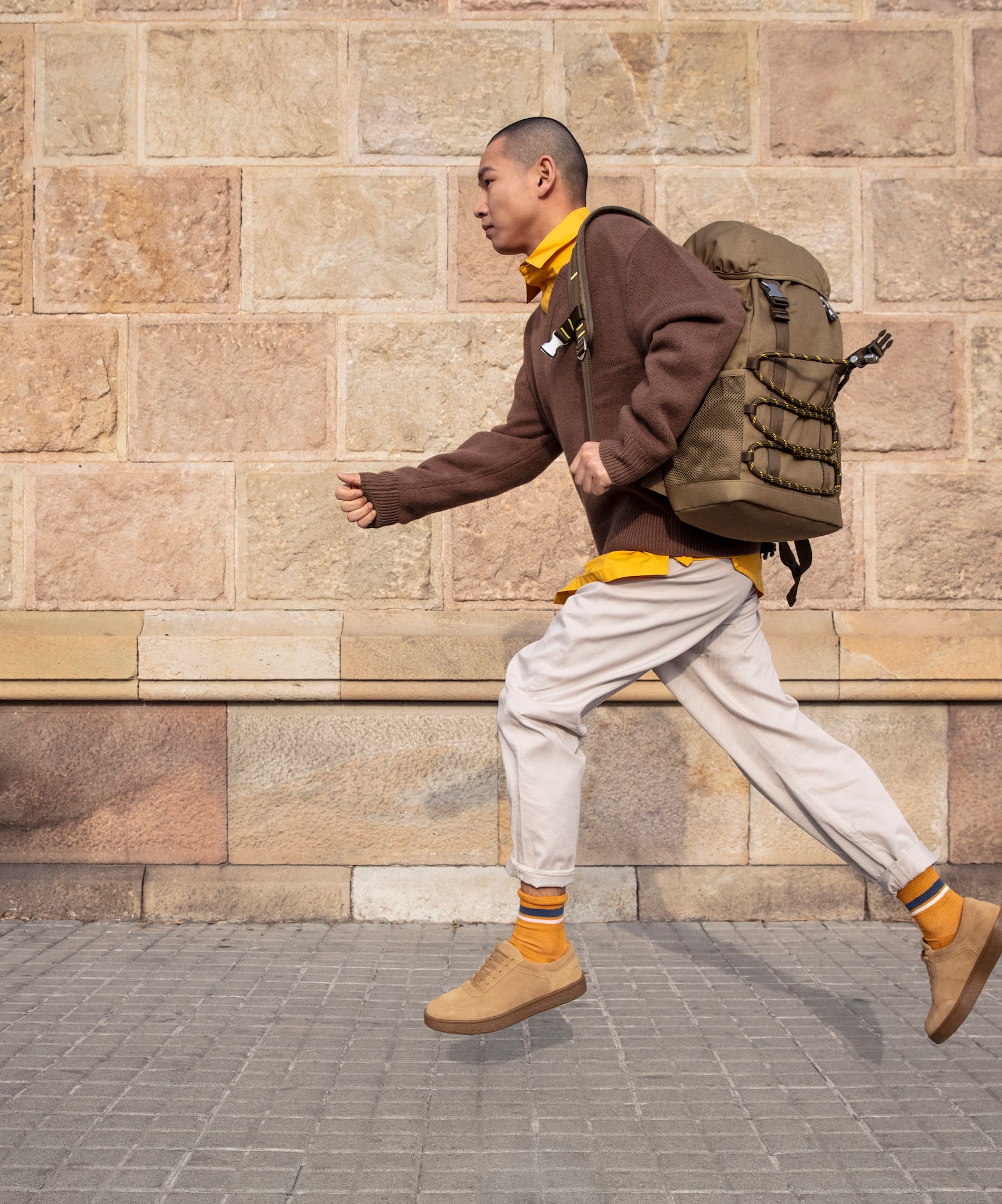 Hombre corriendo con mochila oliva, suéter marrón y zapatillas camel en calle.