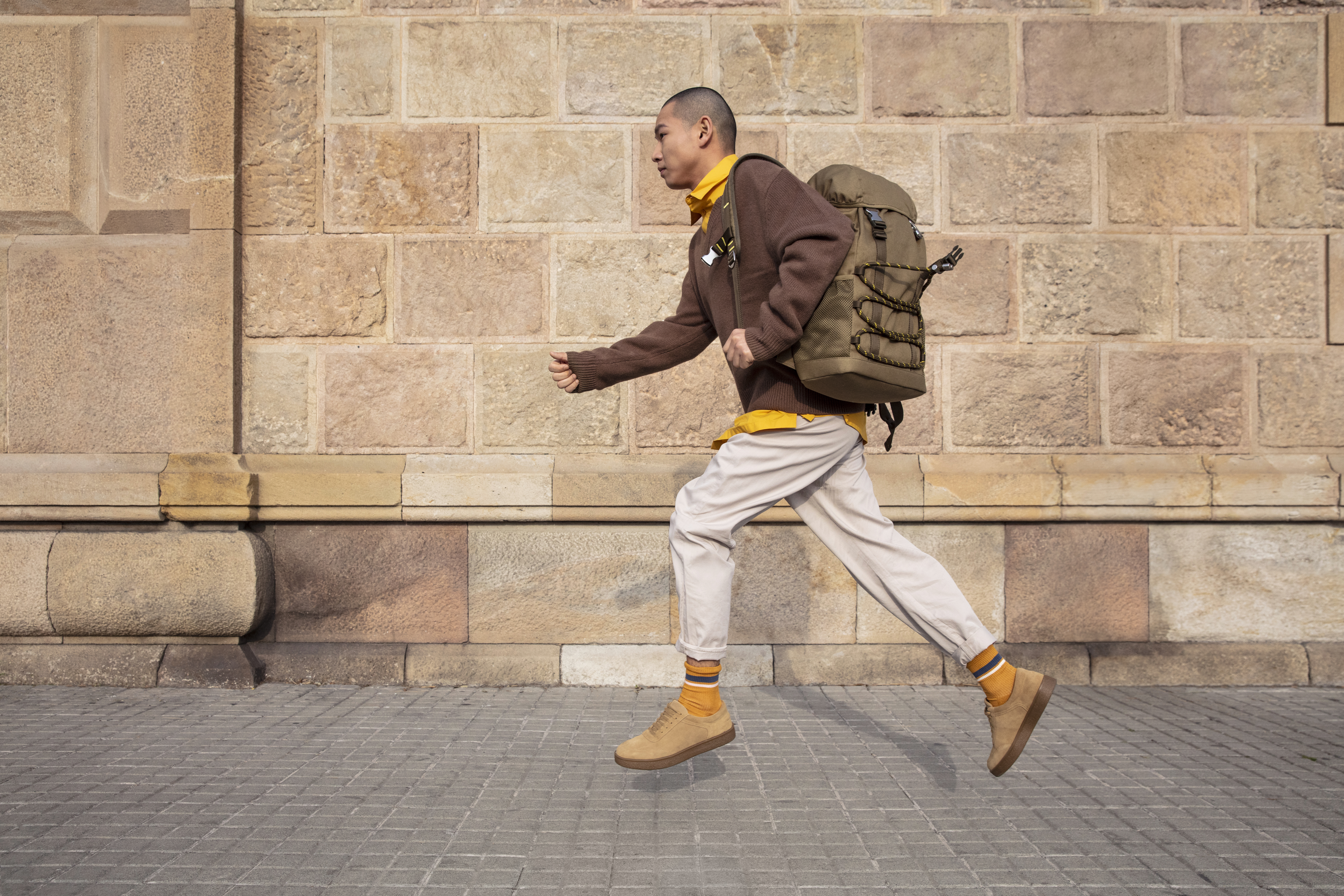 Hombre corriendo con mochila oliva, suéter marrón y zapatillas camel en calle.