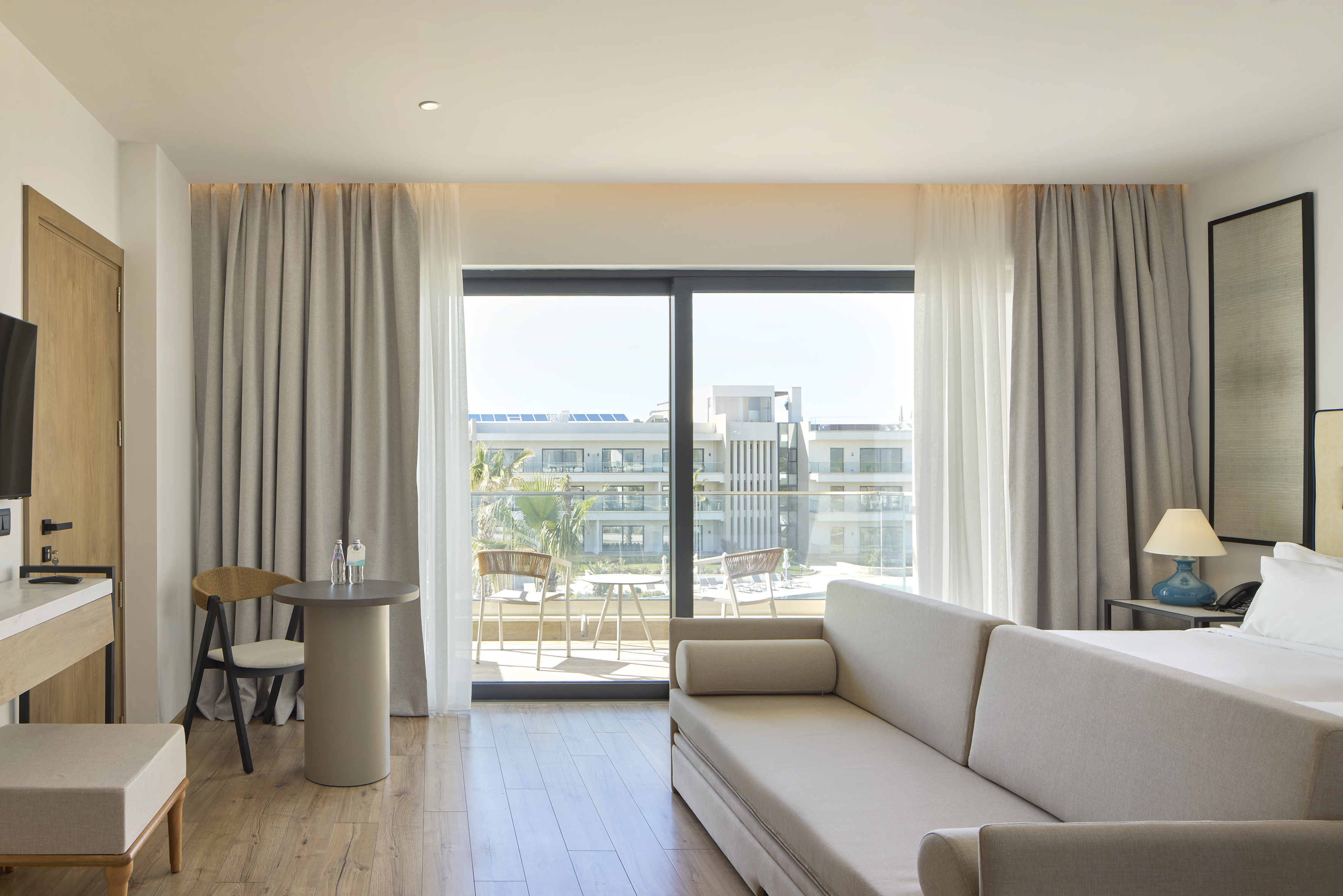 Modern hotel suite with sofa, private balcony overlooking resort buildings and palms.