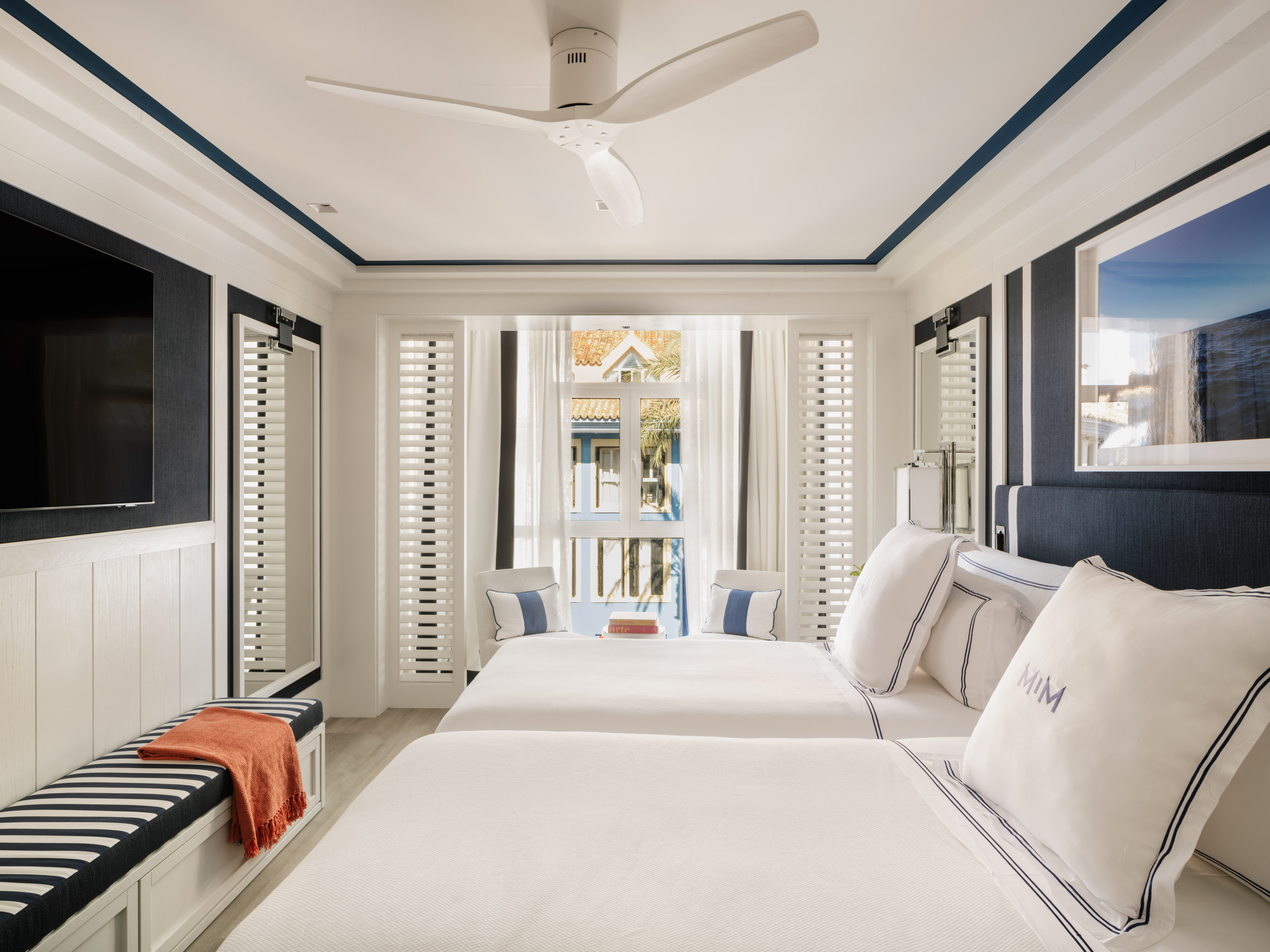 Modern coastal bedroom with two beds, blue accents, white shutters, and ceiling fan.