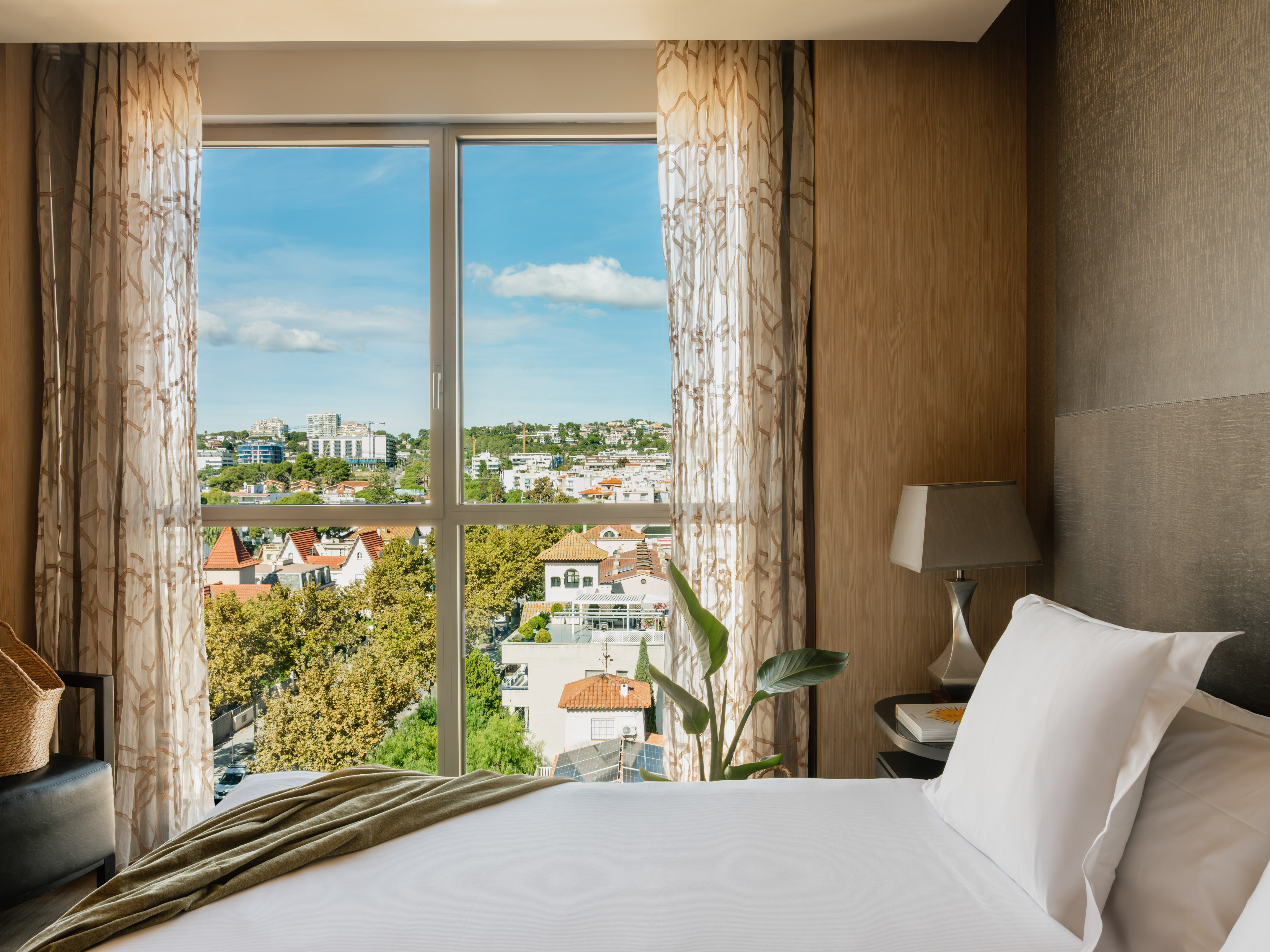Bright bedroom with city view from window, white bed and plant.