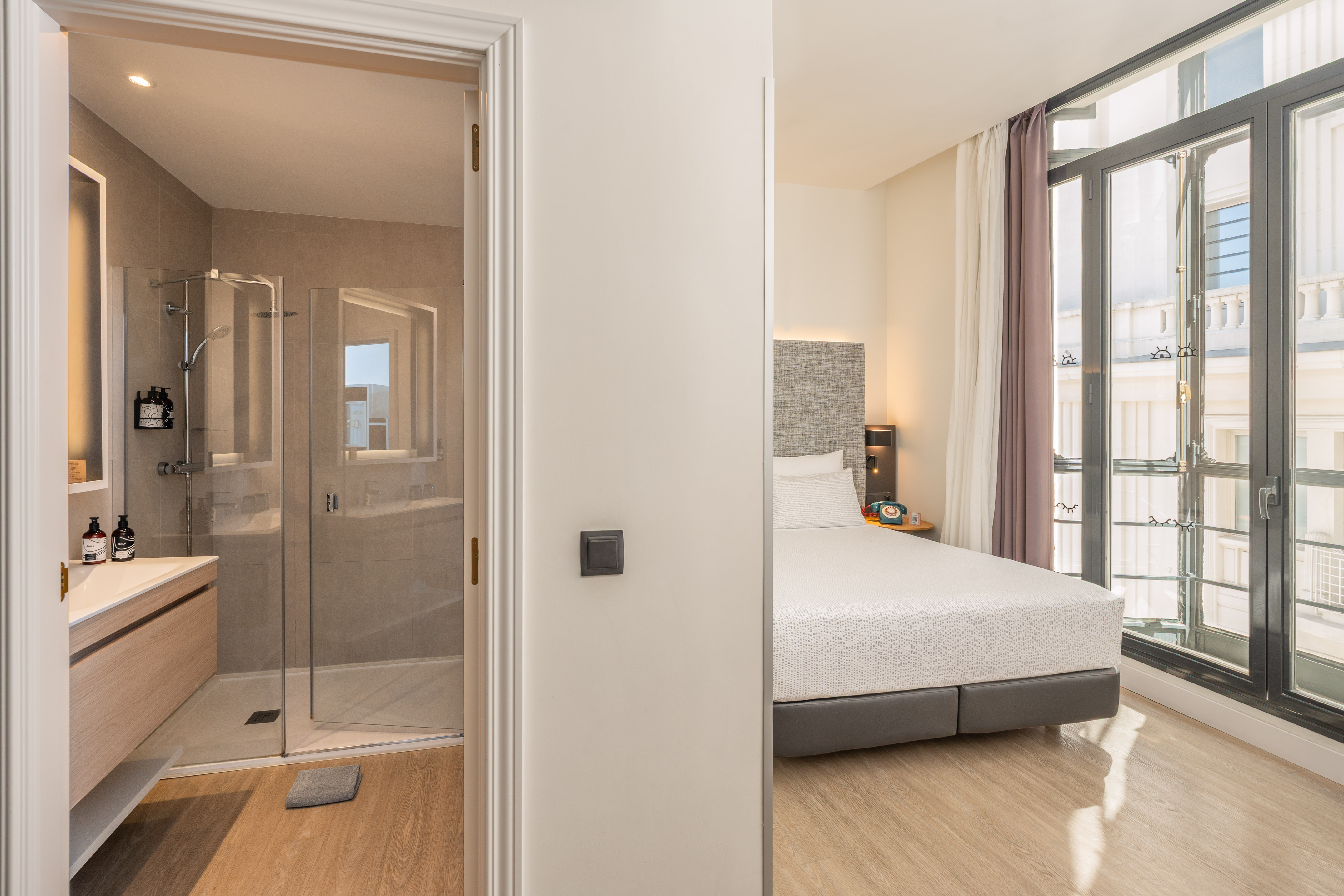 Modern hotel room with bed, large windows, and glass shower bathroom.