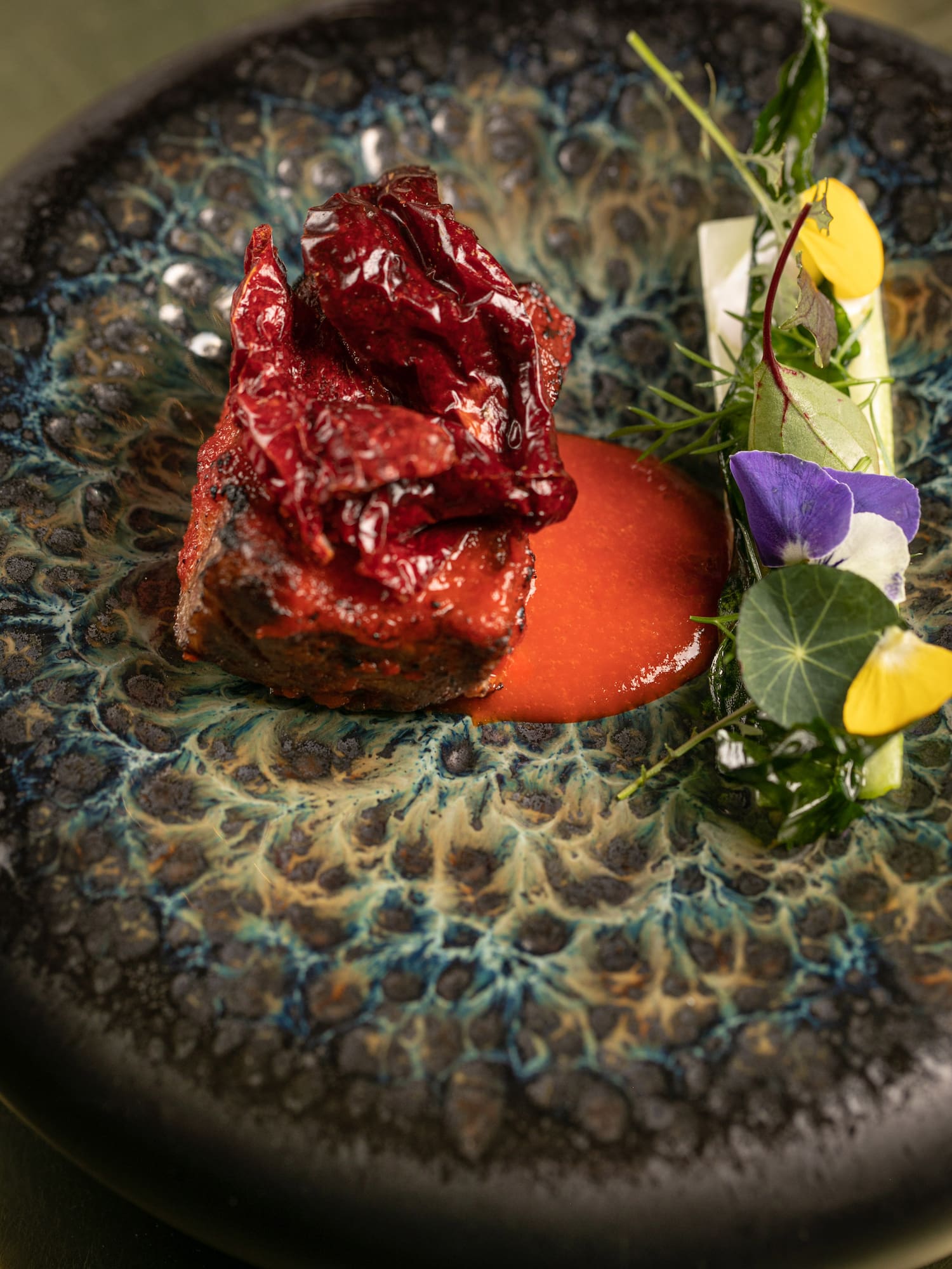 Red chili meat with vibrant sauce, herbs, and edible flowers on a textured plate.