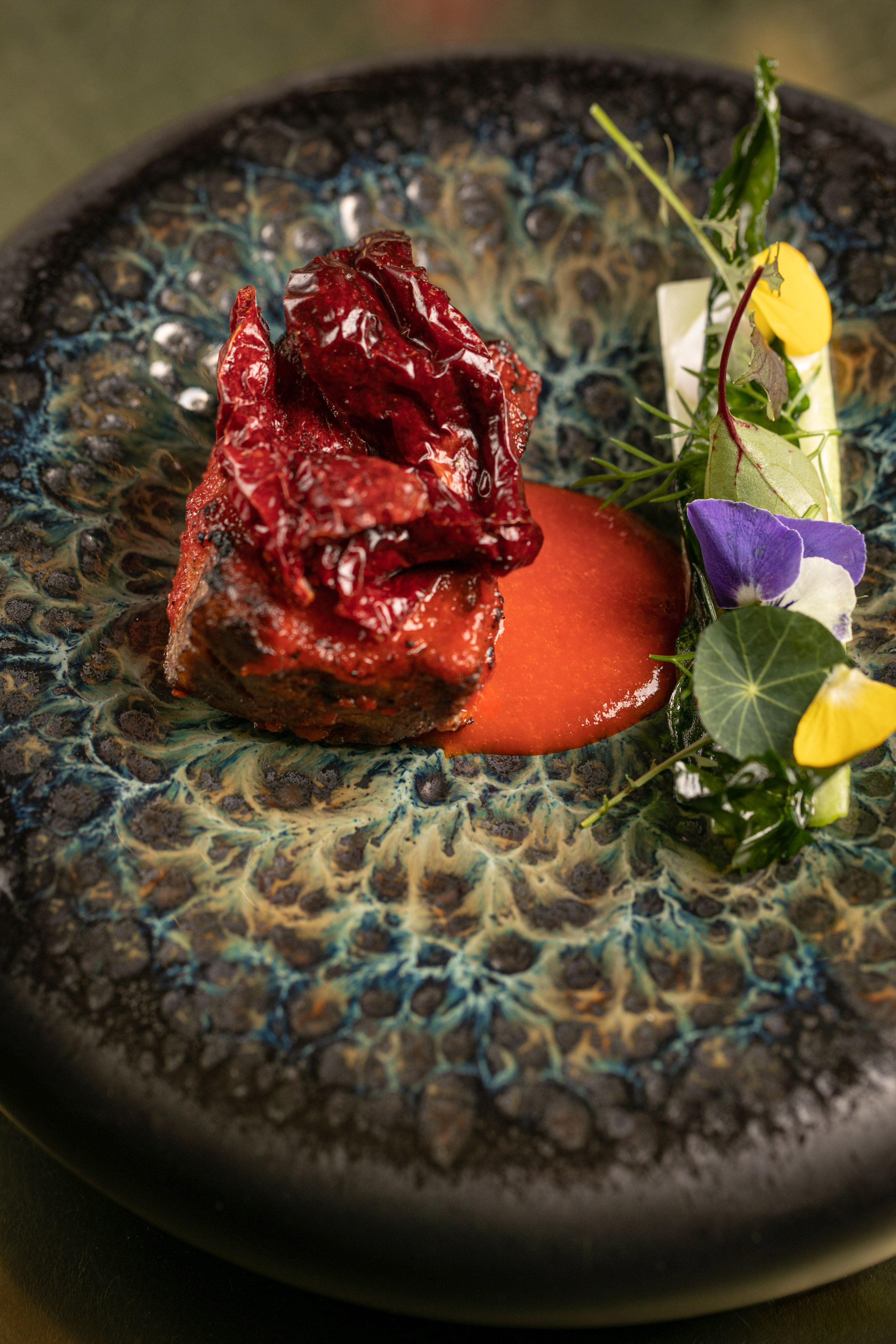 Red chili meat with vibrant sauce, herbs, and edible flowers on a textured plate.