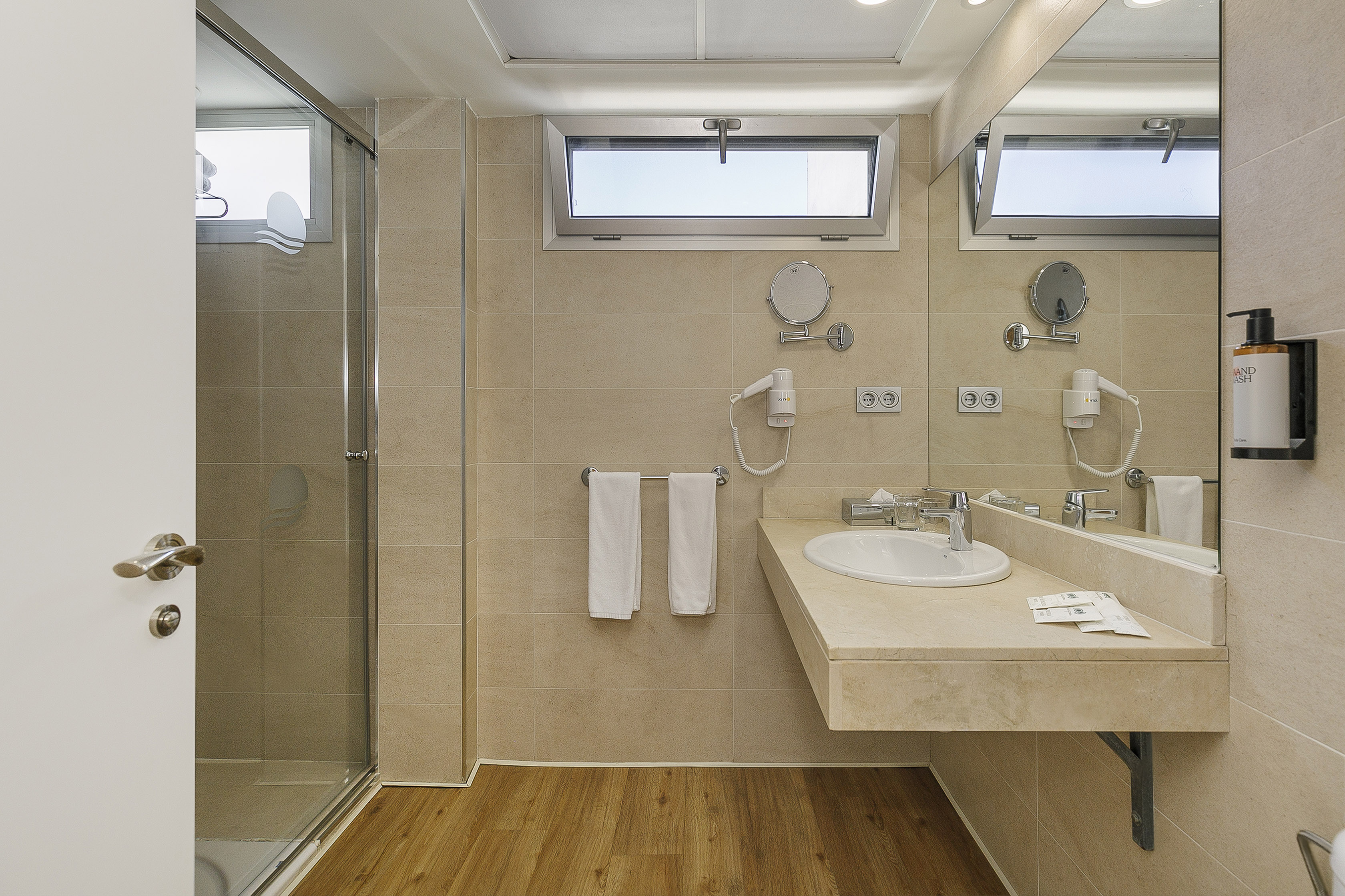 Modern bathroom with glass shower, beige tiled walls, wood floor, and vanity.