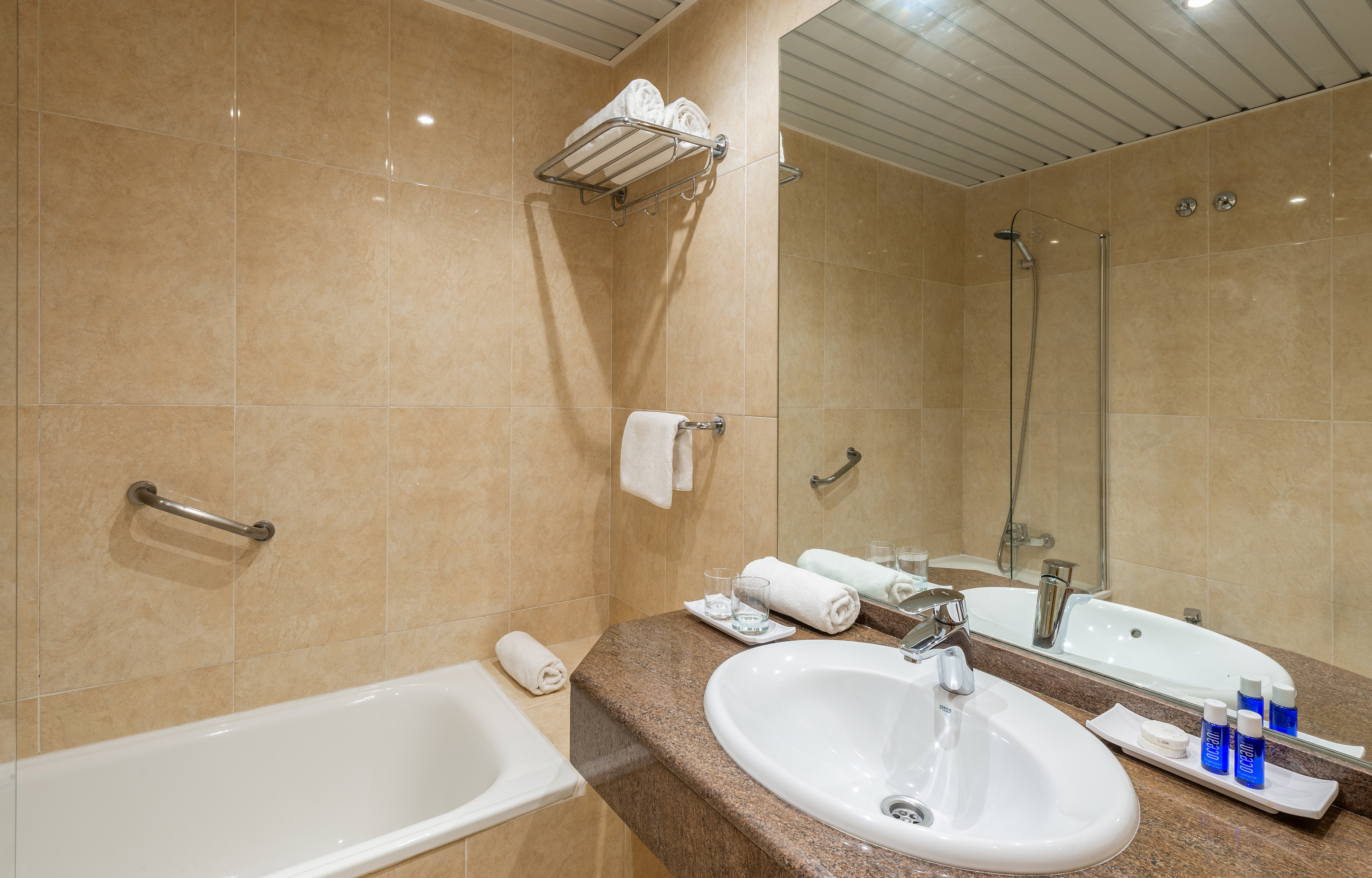 Hotel bathroom with bathtub, vanity sink, mirror, beige tiles, and towels.