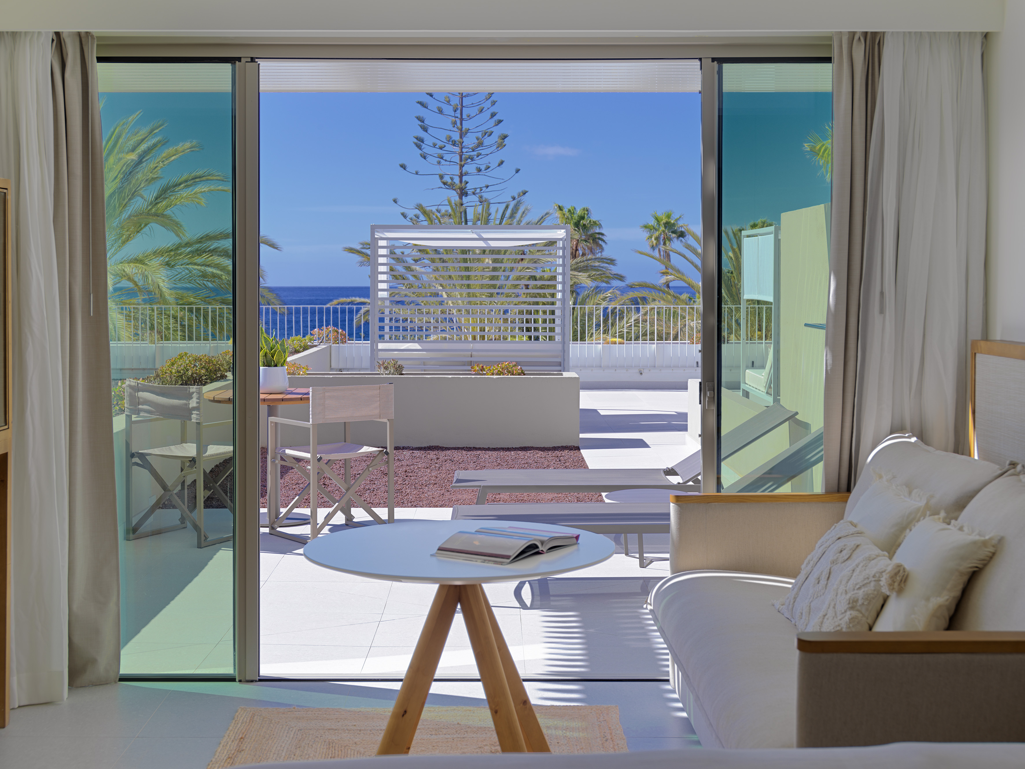 Modern ocean view hotel room with open terrace, sofa, and palm trees.