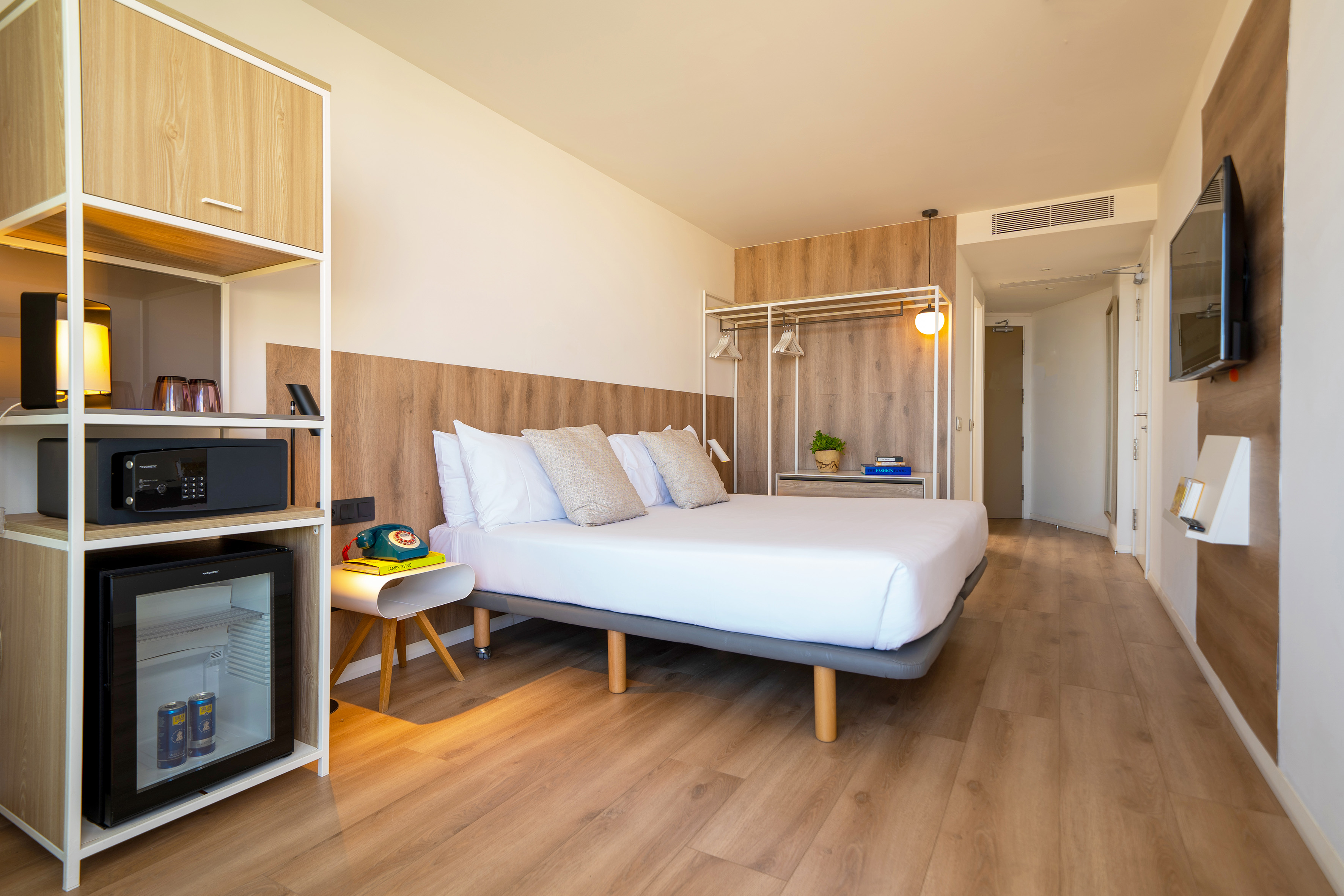 Modern hotel room with double bed, wood floor, mini-fridge, safe, and open wardrobe.