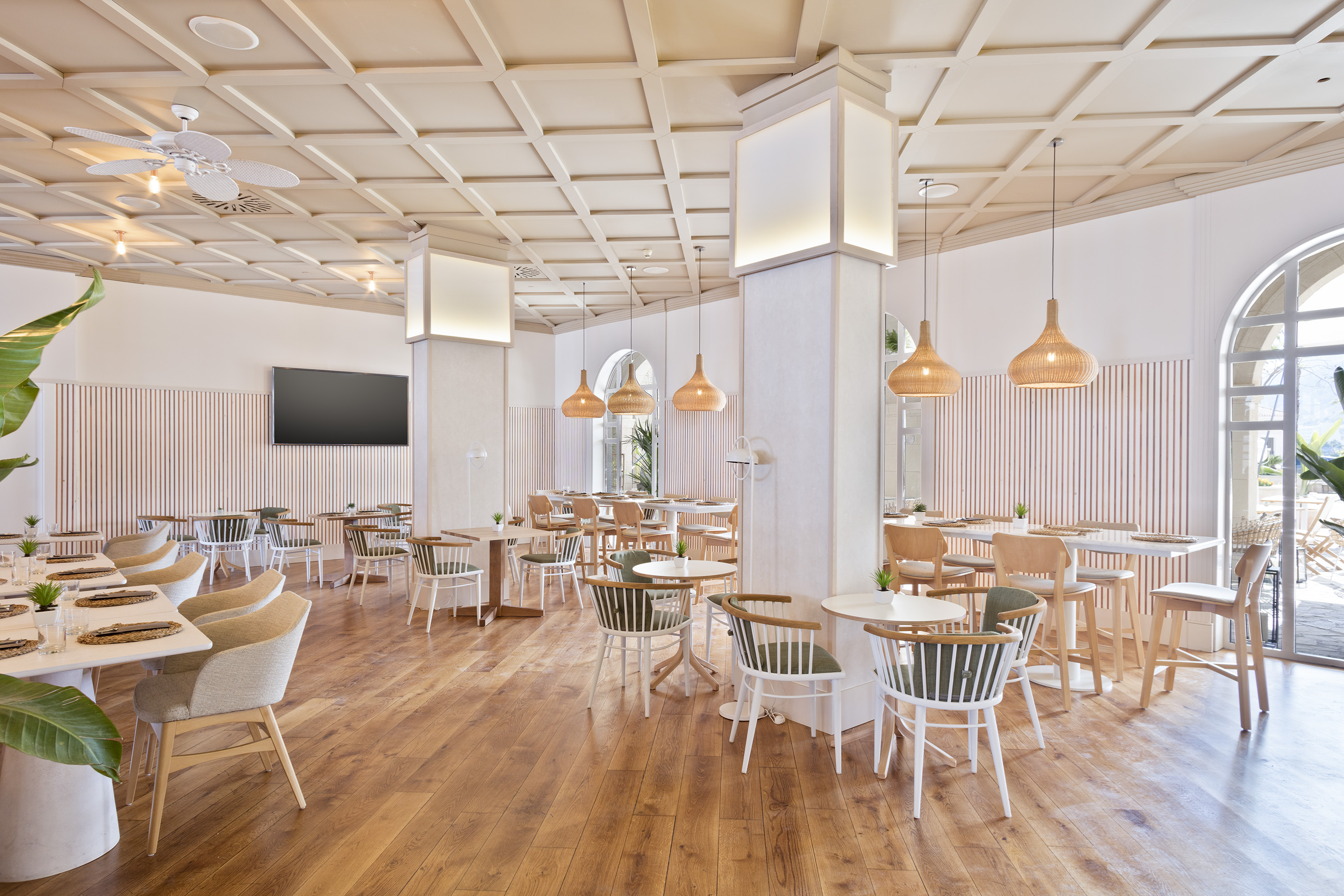 Modern restaurant dining room with wooden floor, light furniture, and woven lamps.
