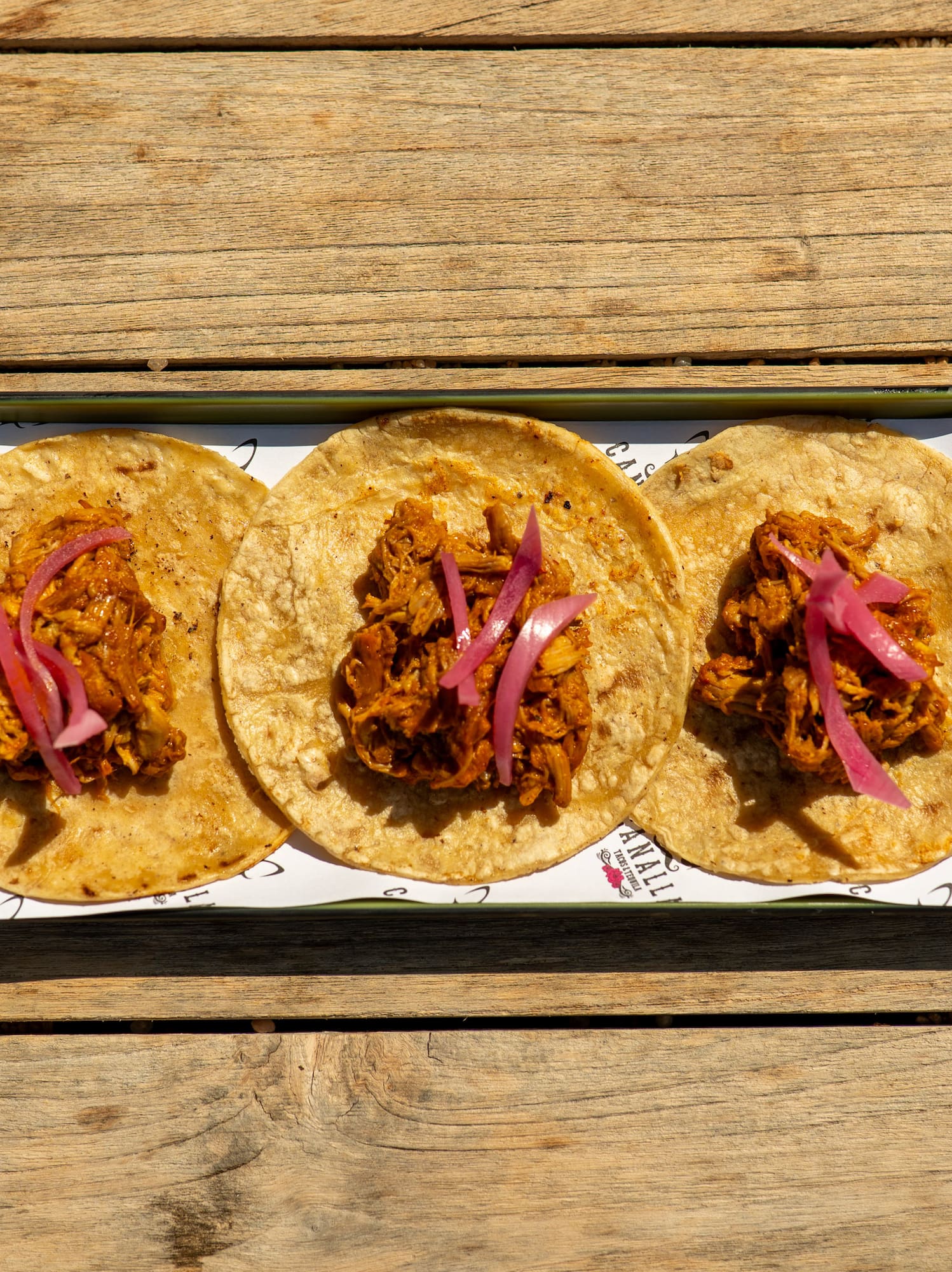 Three cochinita pibil tacos with pickled red onions on a wooden table.
