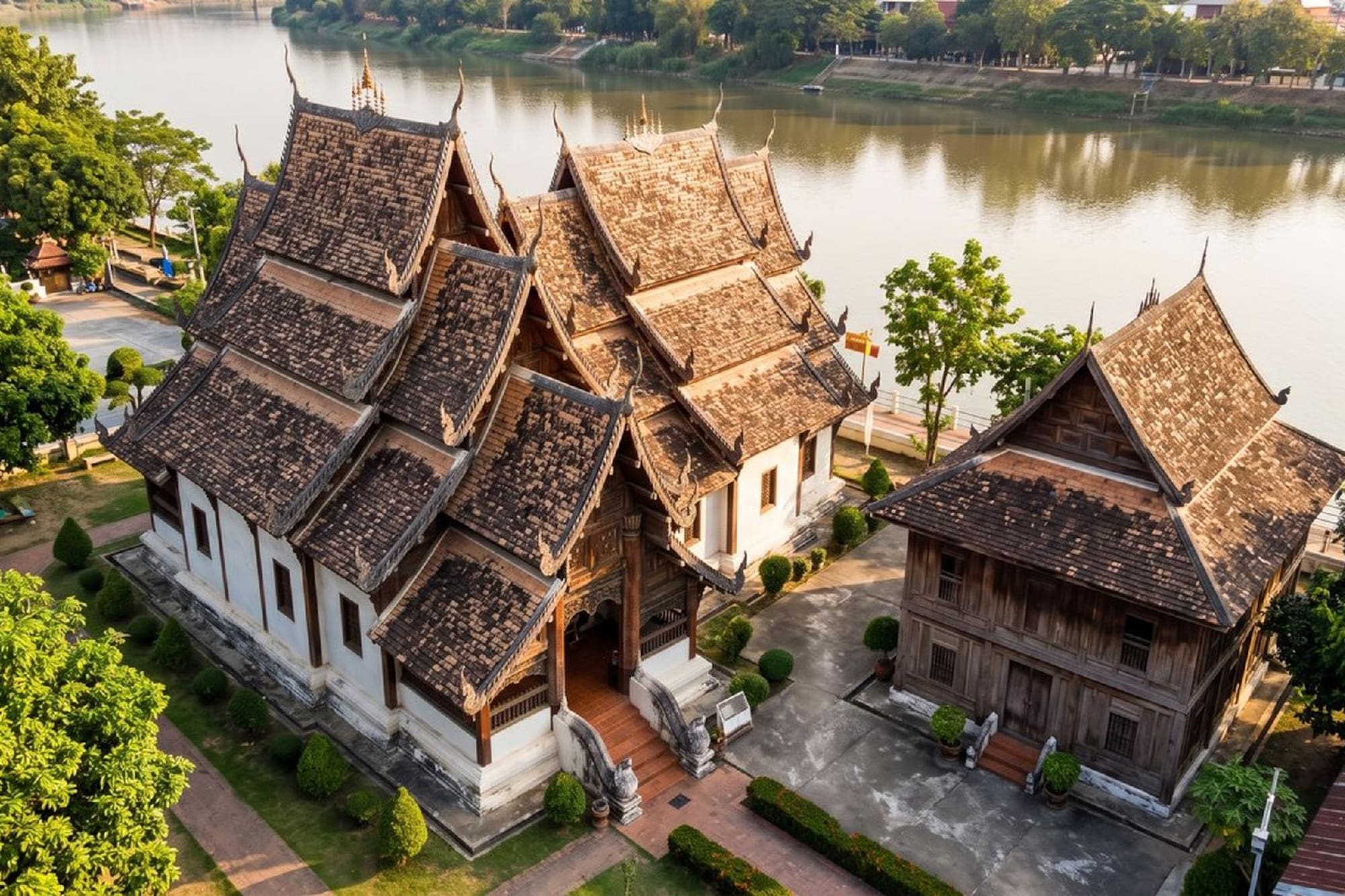 Thai temple complex by river with traditional roofs and wooden buildings.