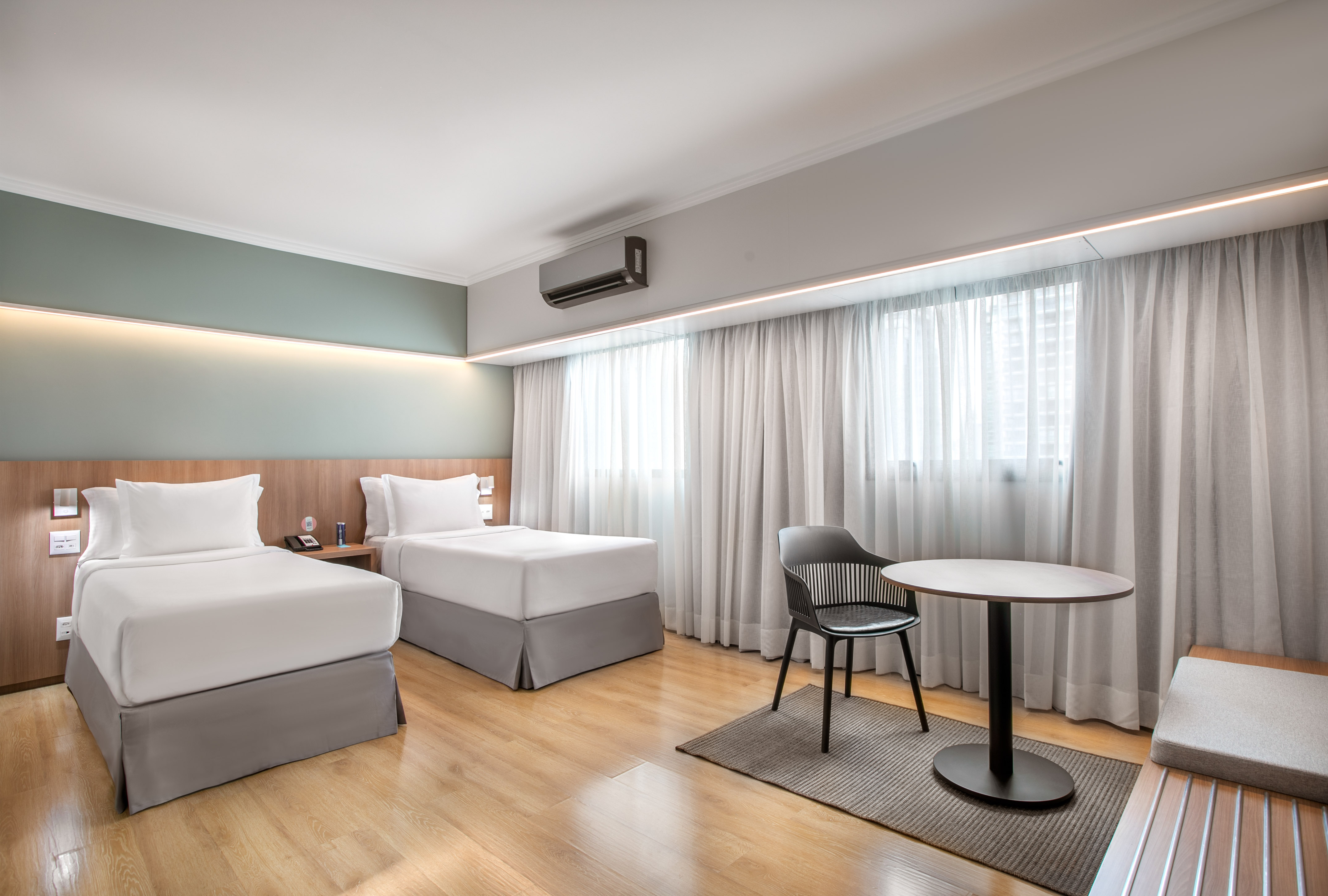 Bright hotel room with two twin beds, wooden floor, and light curtains.
