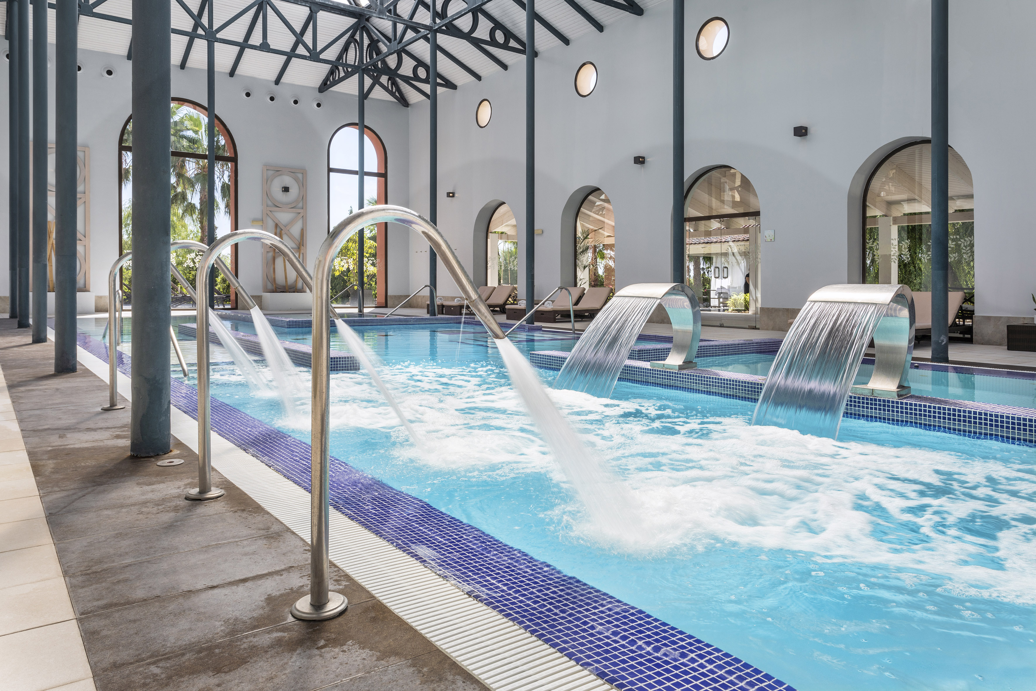 Indoor spa pool with hydrotherapy jets, waterfalls, and arched windows.