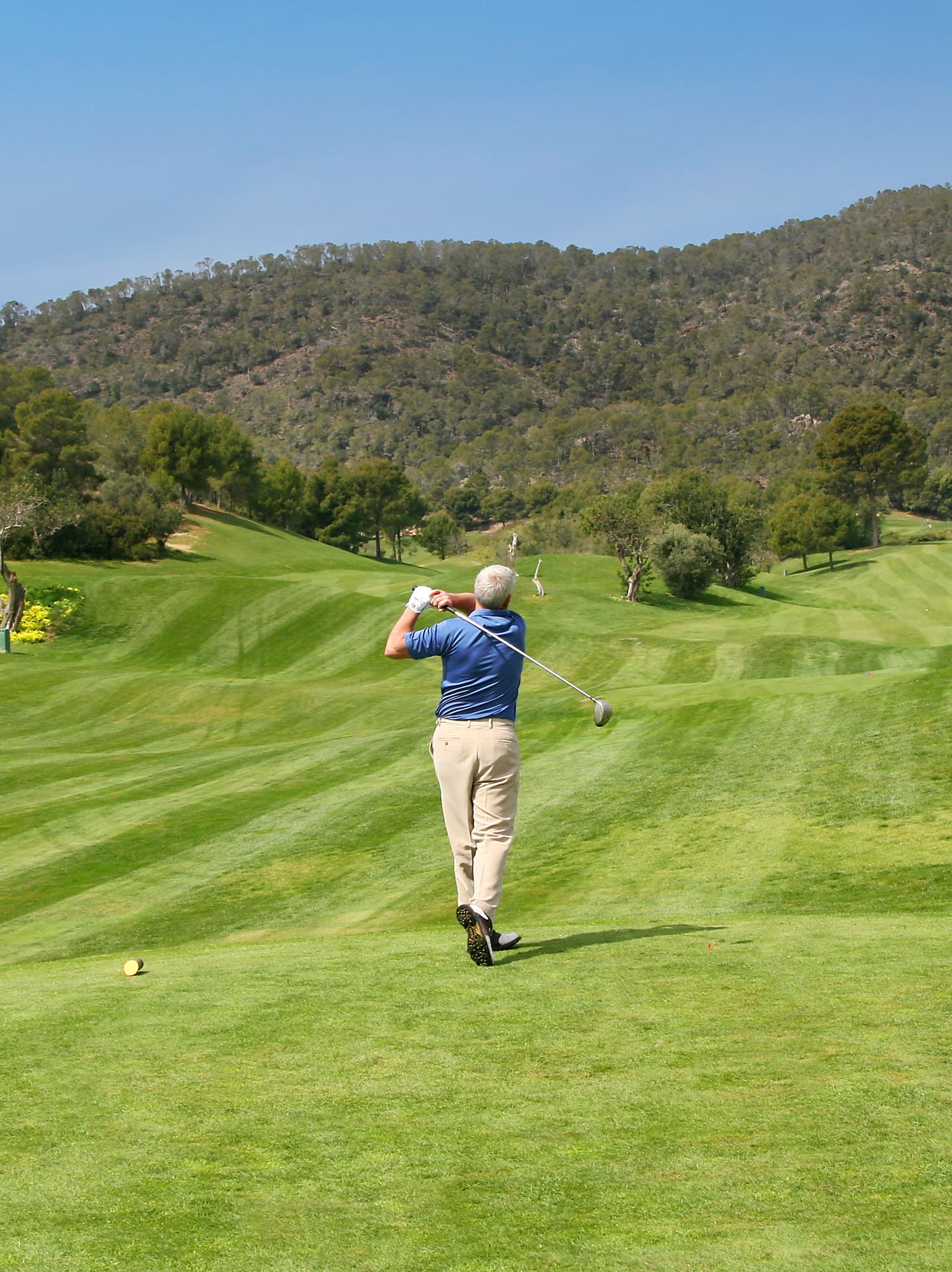 Man in blue shirt golfing, mid-swing, on a green course with hills.