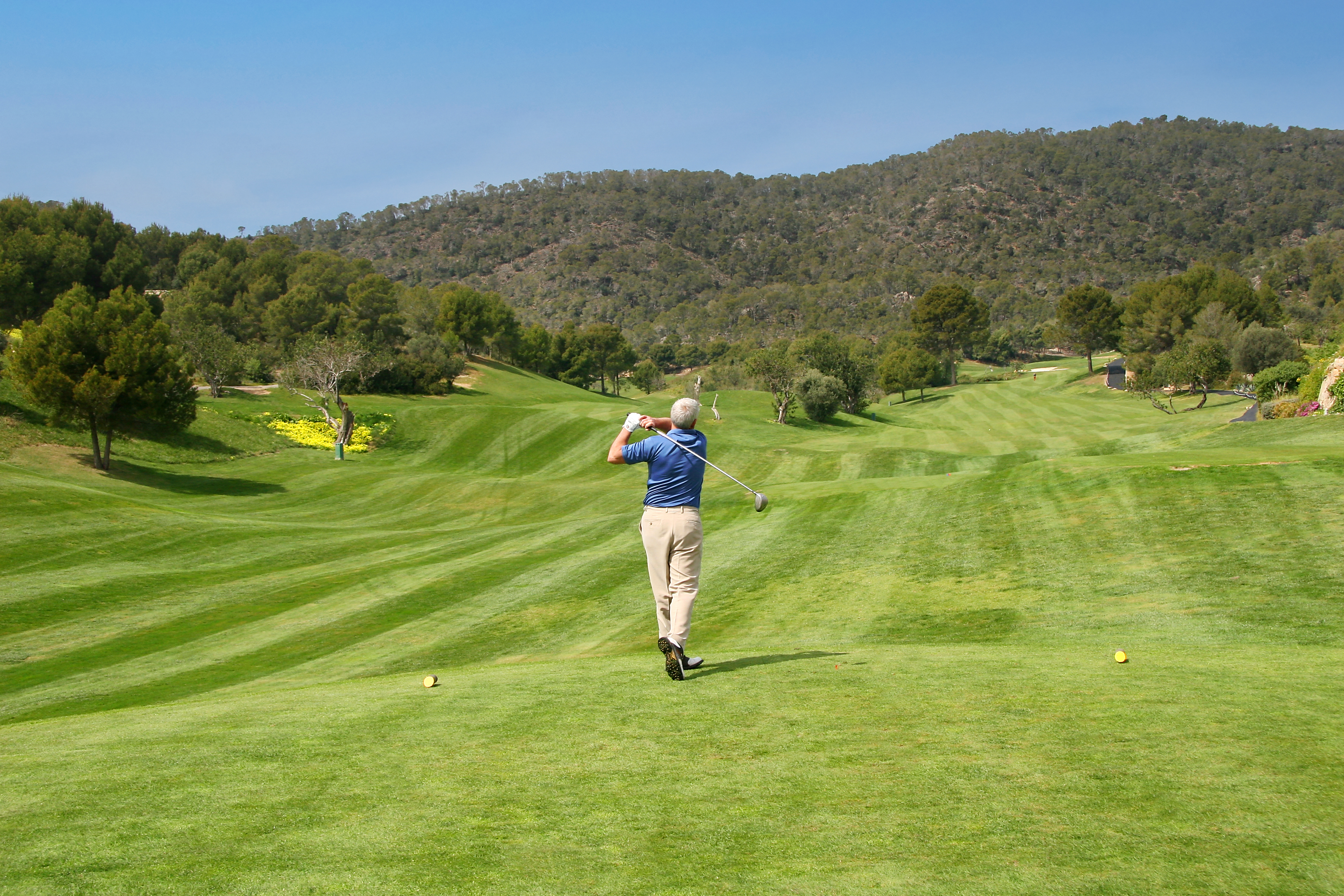 Man in blue shirt golfing, mid-swing, on a green course with hills.