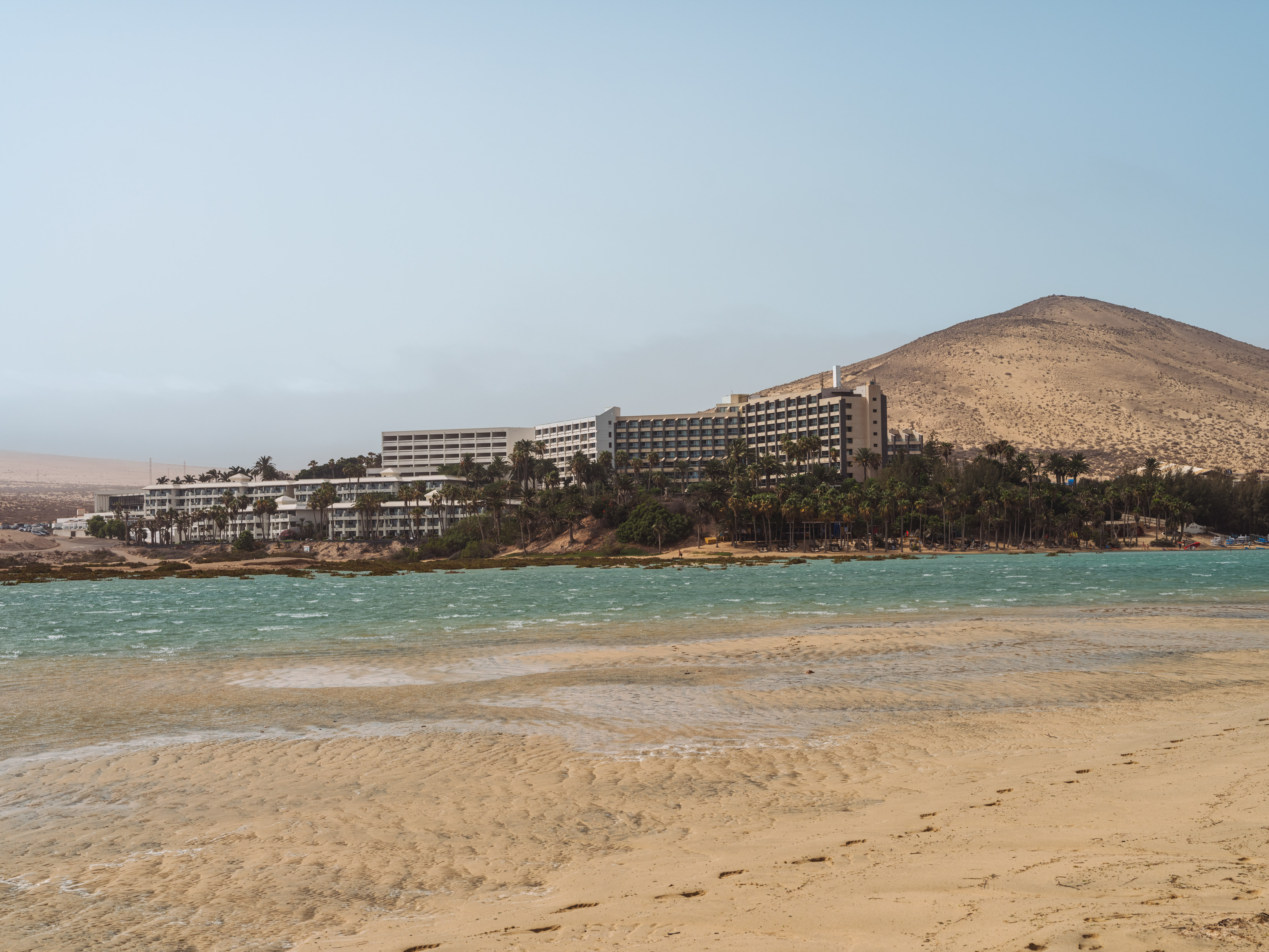 Resort with palm trees, sandy beach, turquoise lagoon, and arid mountain.