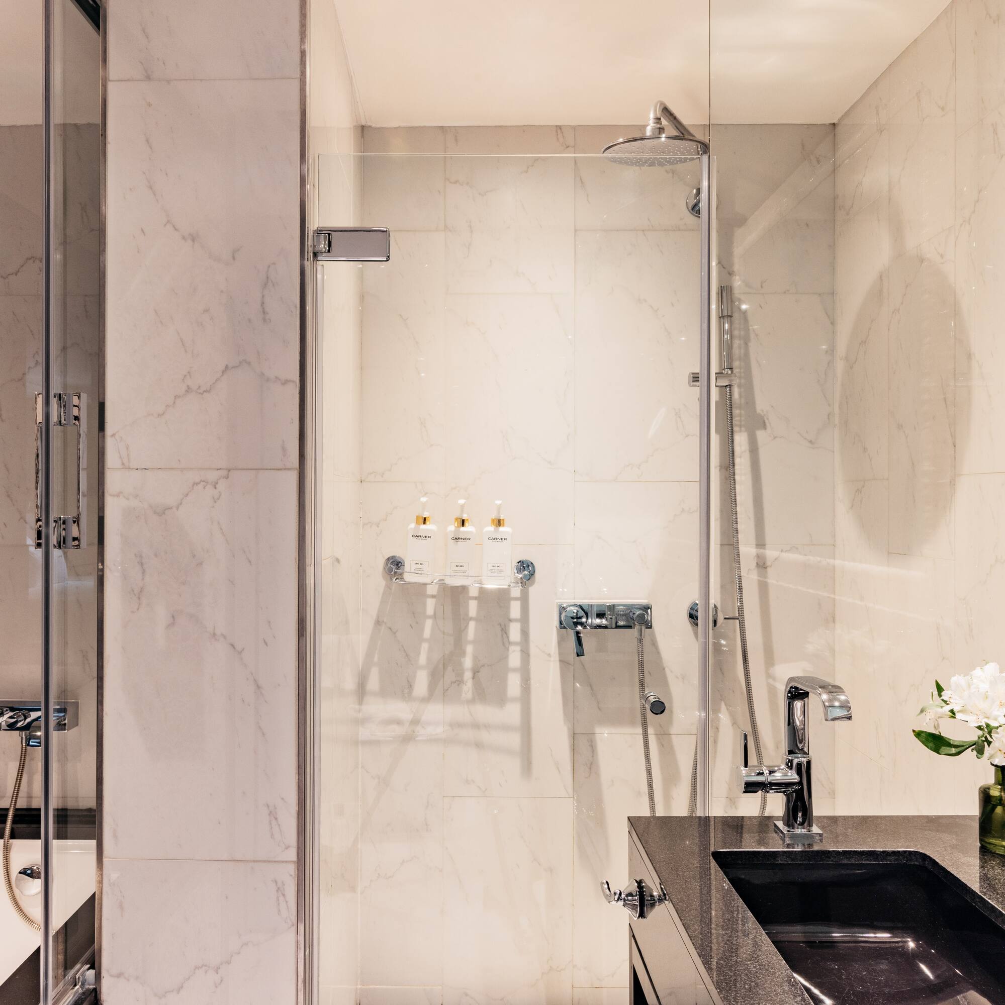 Baño moderno con ducha de cristal, bañera y lavabo negro sobre mármol blanco.