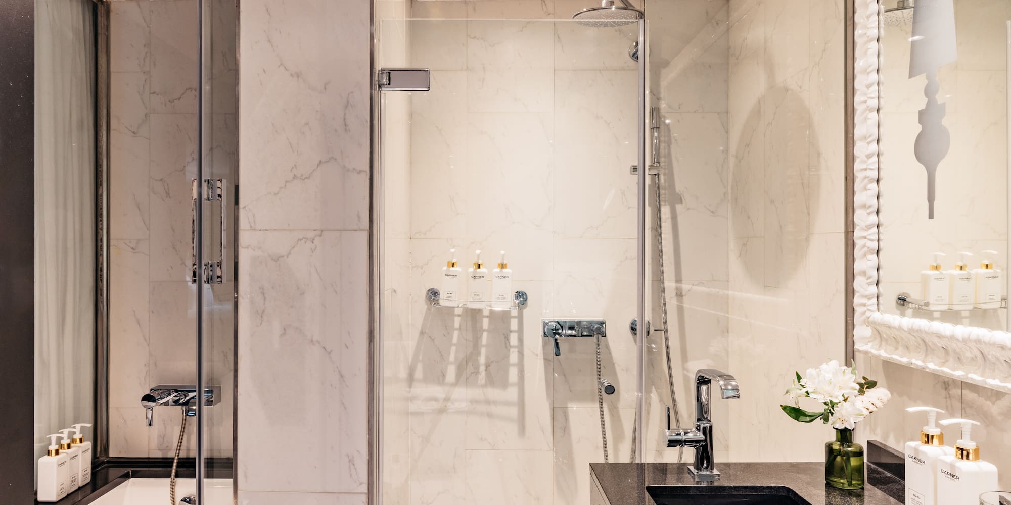 Baño moderno con ducha de cristal, bañera y lavabo negro sobre mármol blanco.