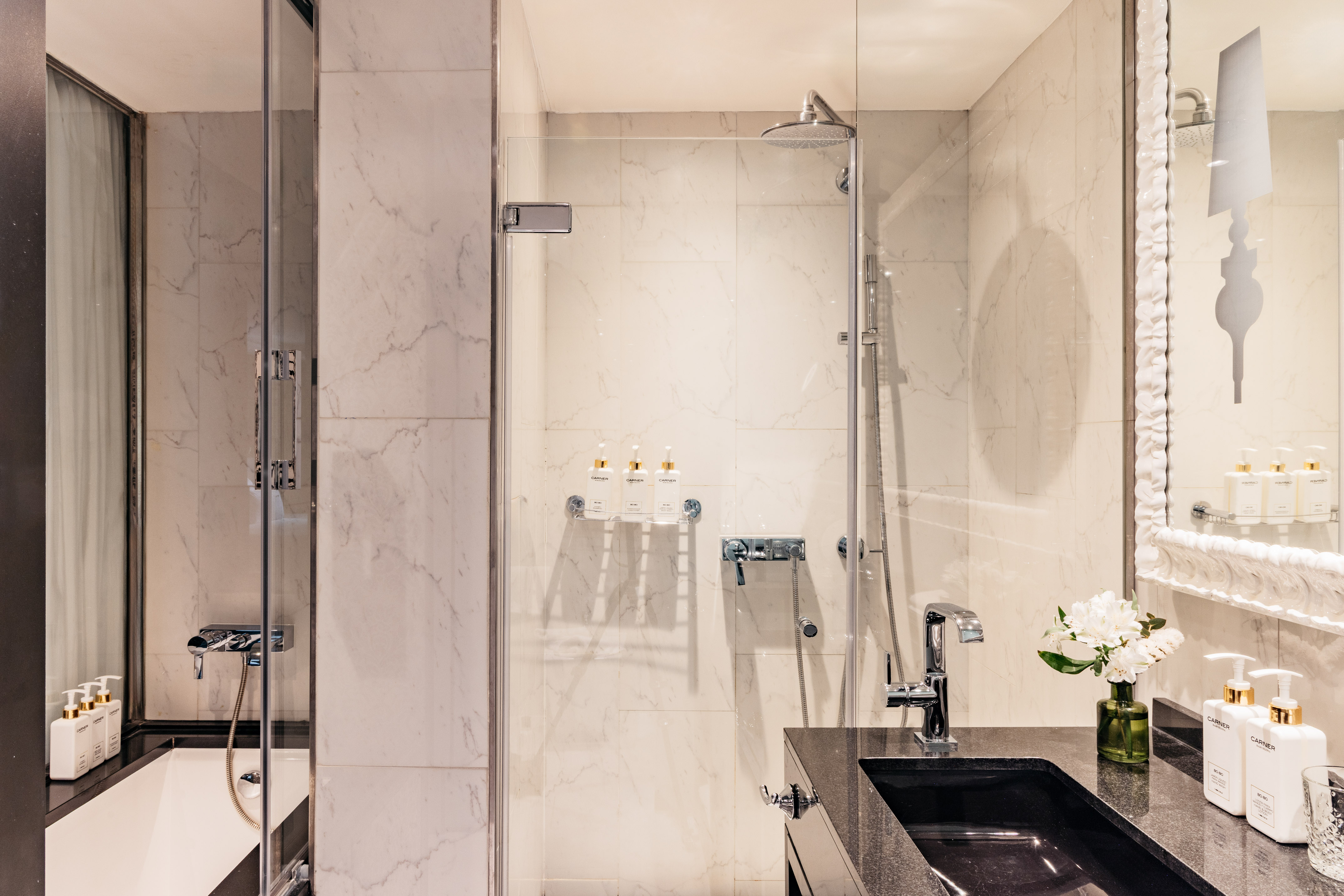 Baño moderno con ducha de cristal, bañera y lavabo negro sobre mármol blanco.