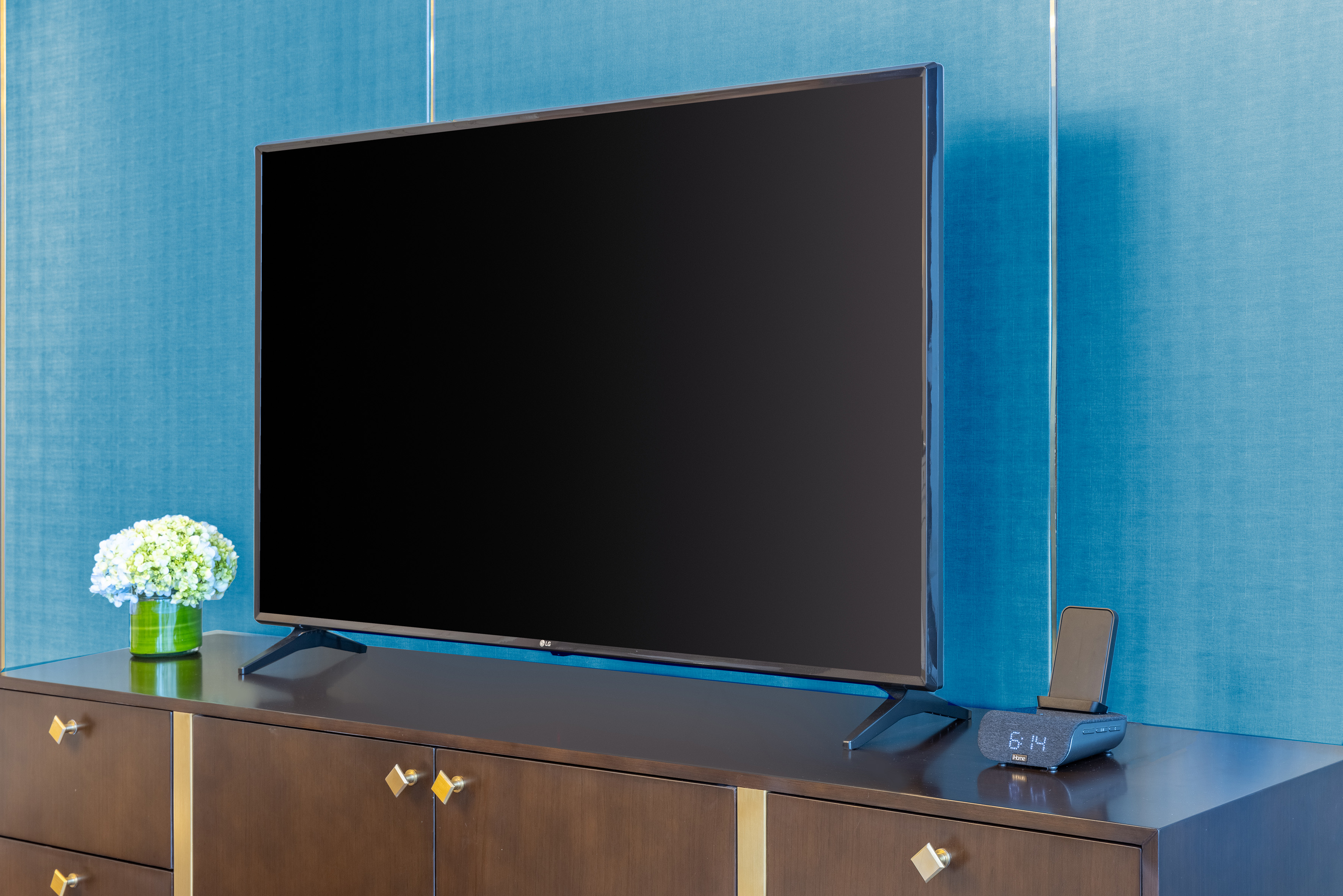 LG TV on a dark dresser with blue wall, flowers, and digital clock.