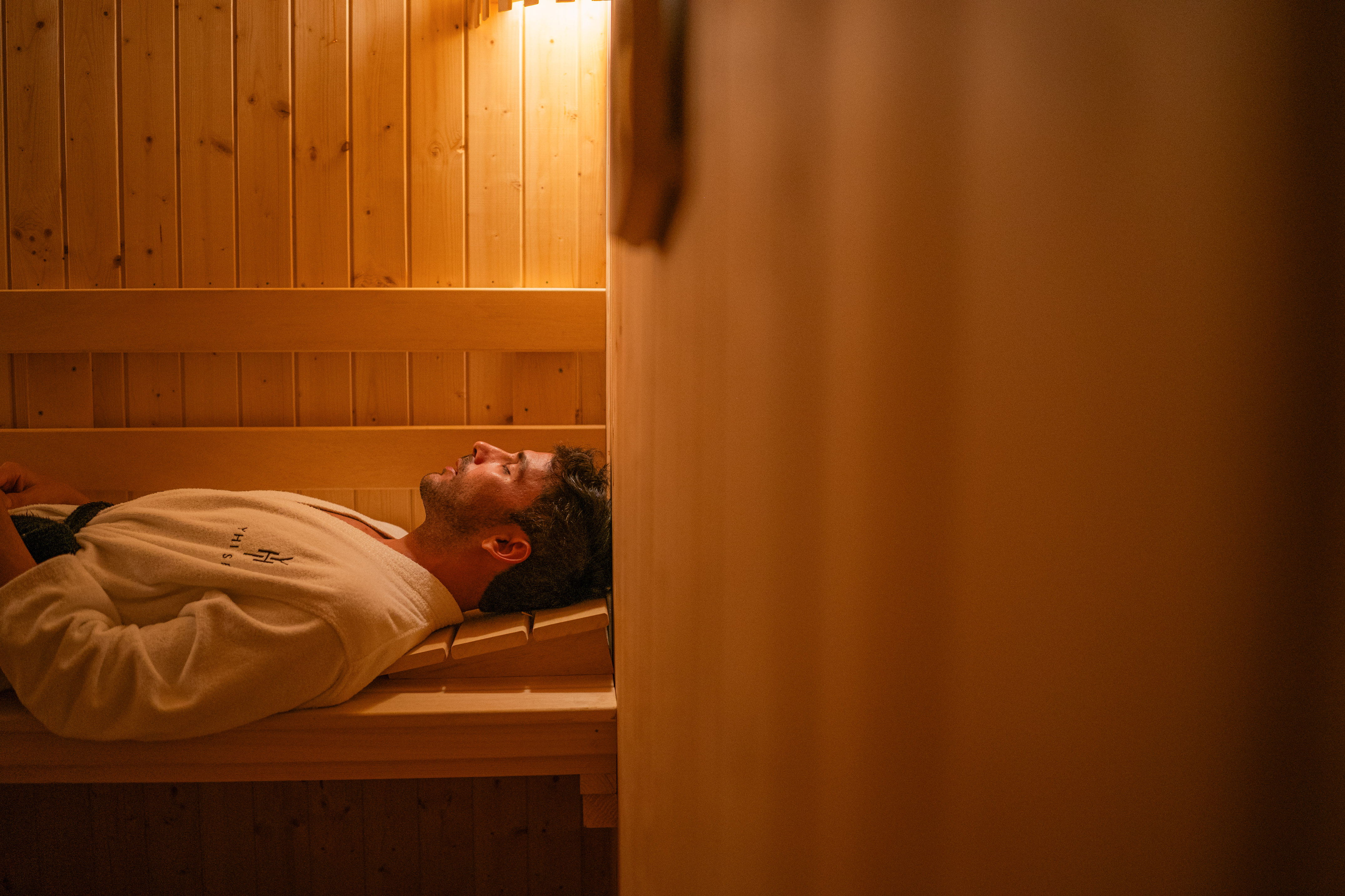 Relaxed man in white bathrobe lying on wooden bench in sauna.
