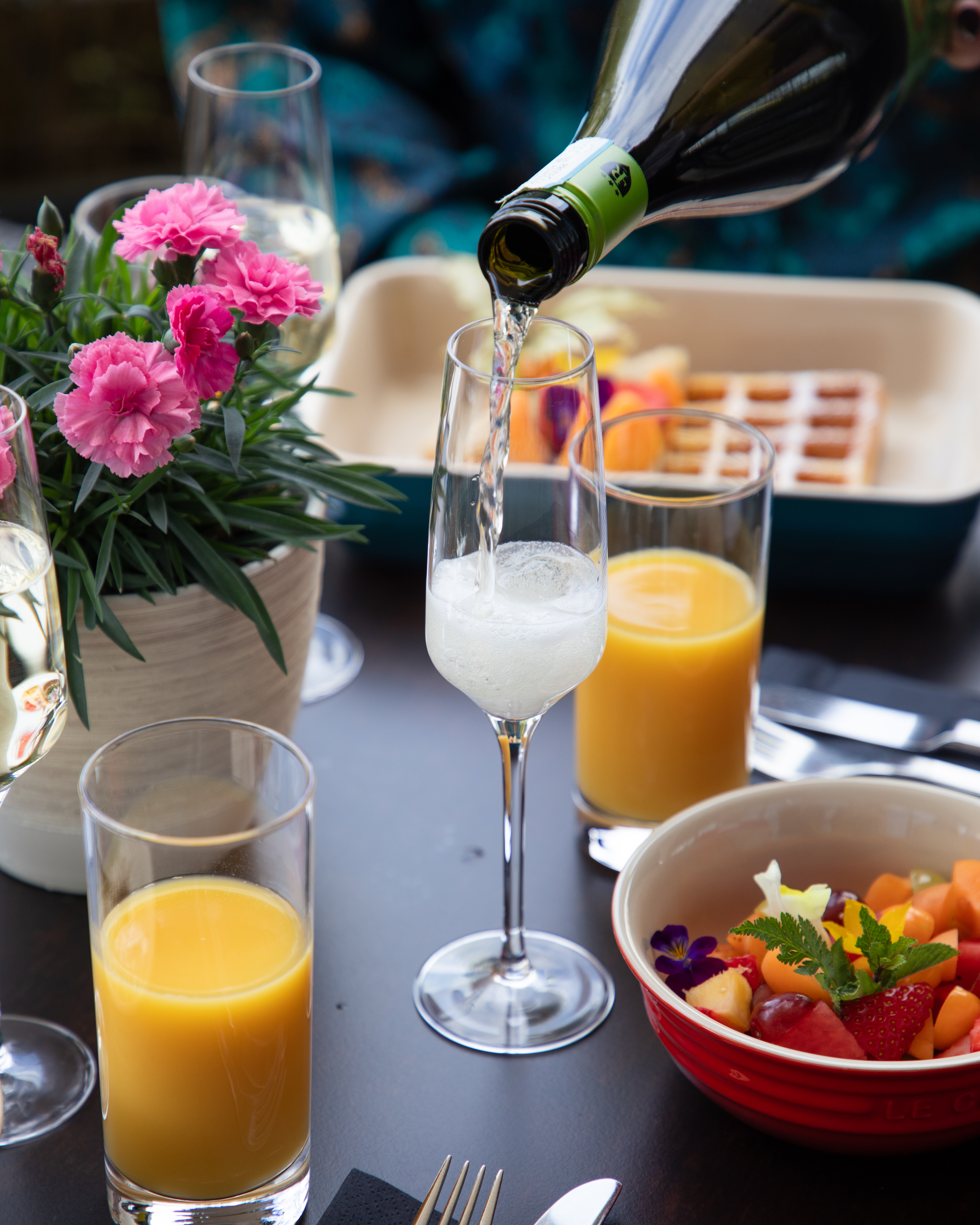 Pouring sparkling wine at brunch with fruit salad, orange juice, and pink flowers.