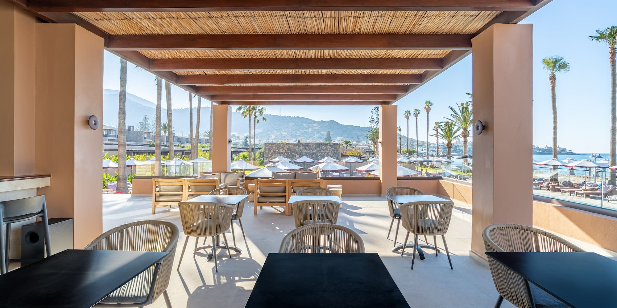 Resort outdoor dining area with bamboo ceiling, beach, palm trees, and mountain view.