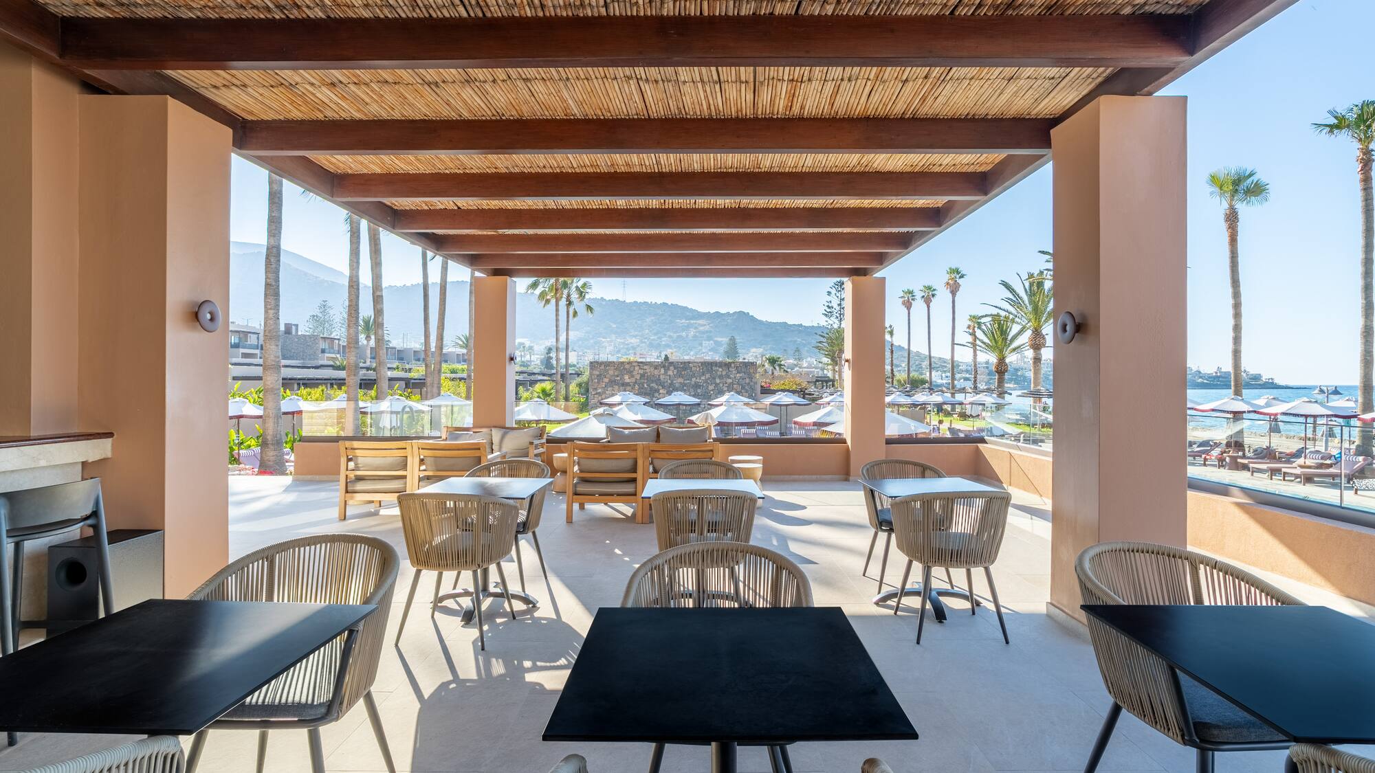 Resort outdoor dining area with bamboo ceiling, beach, palm trees, and mountain view.