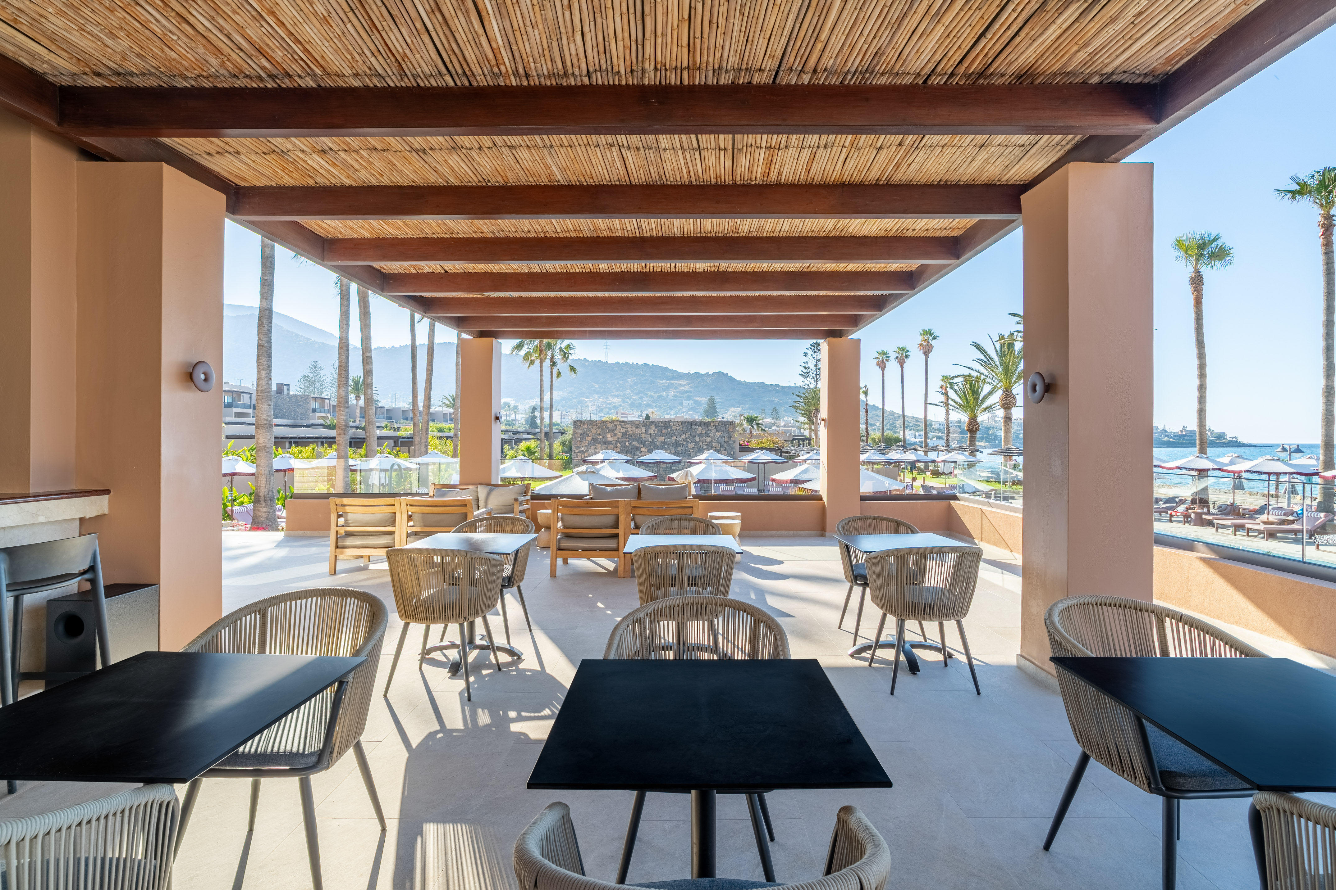 Resort outdoor dining area with bamboo ceiling, beach, palm trees, and mountain view.
