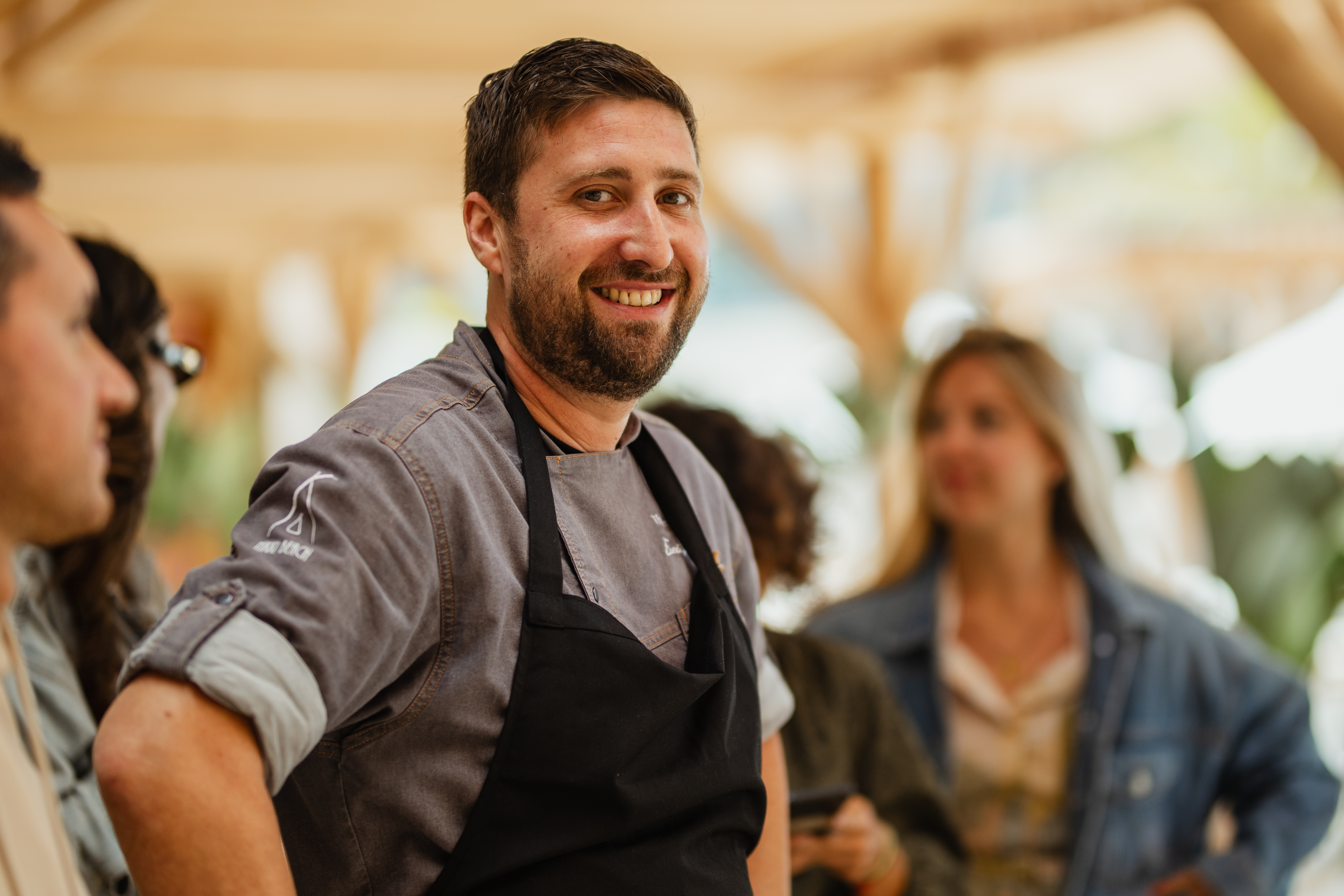 Smiling bearded chef in uniform and black apron at a culinary event.