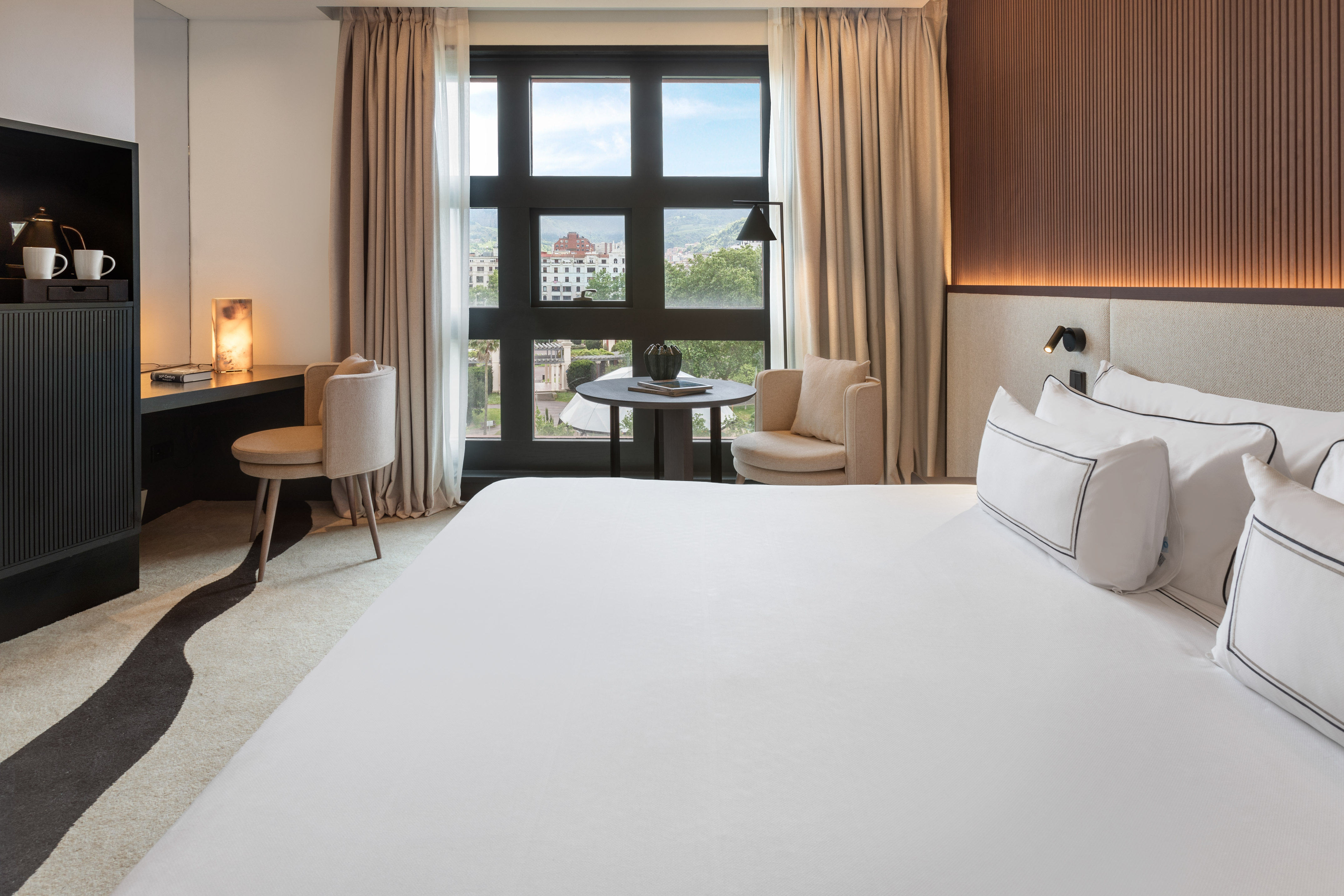 Modern hotel bedroom with white bed, desk, and city view from large window.
