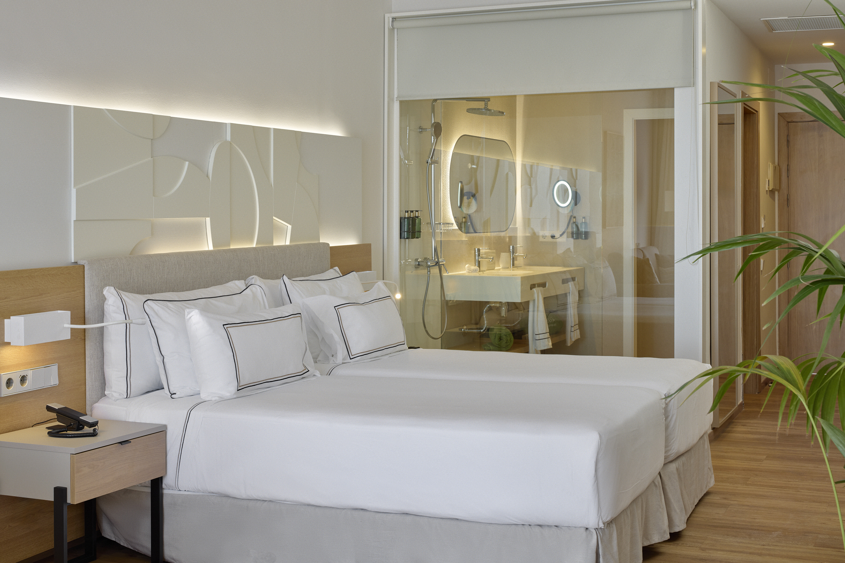Modern hotel room with two single beds, illuminated headboard, and glass bathroom.