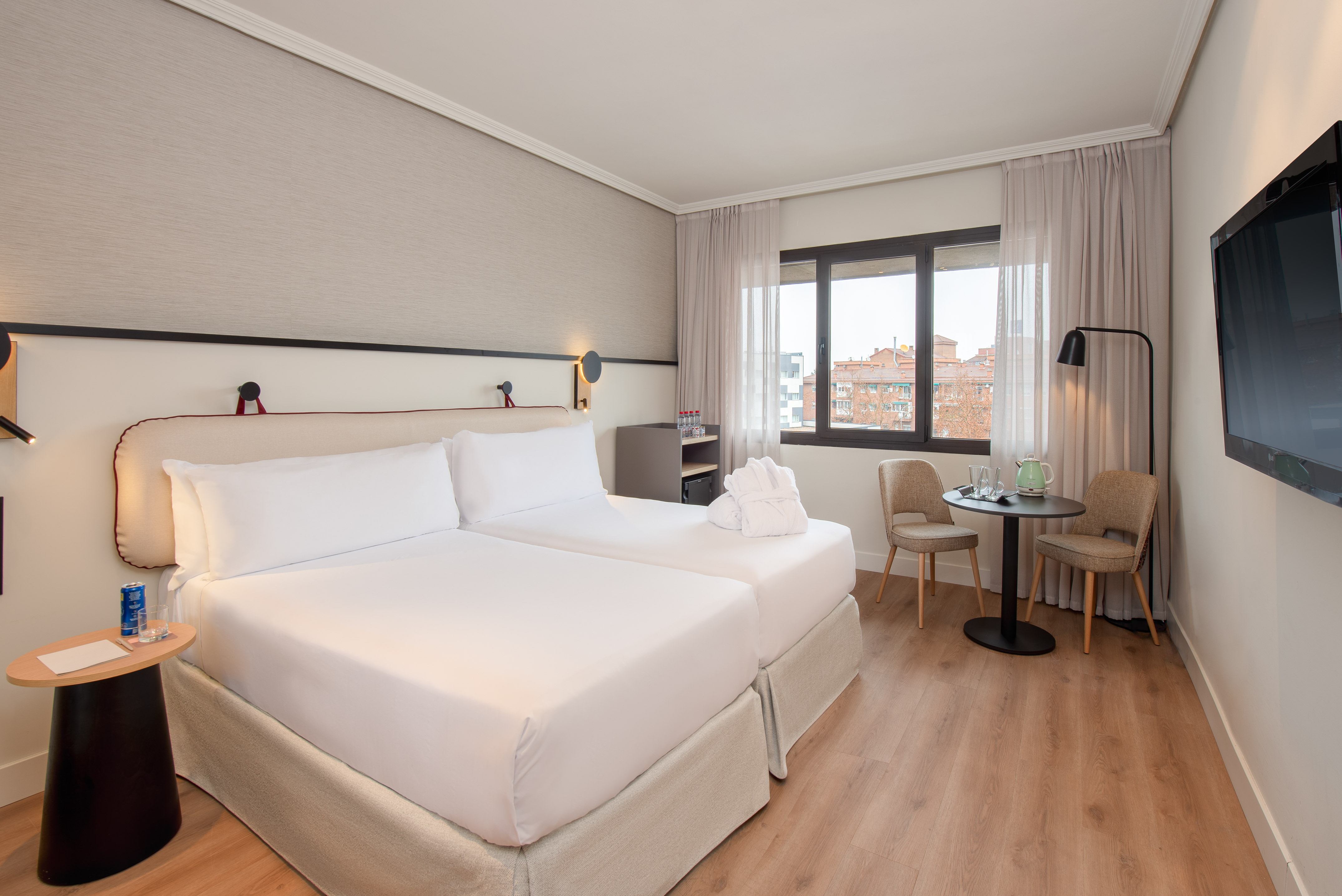 Bright modern hotel room with two twin beds, wood floor, window, and sitting area.
