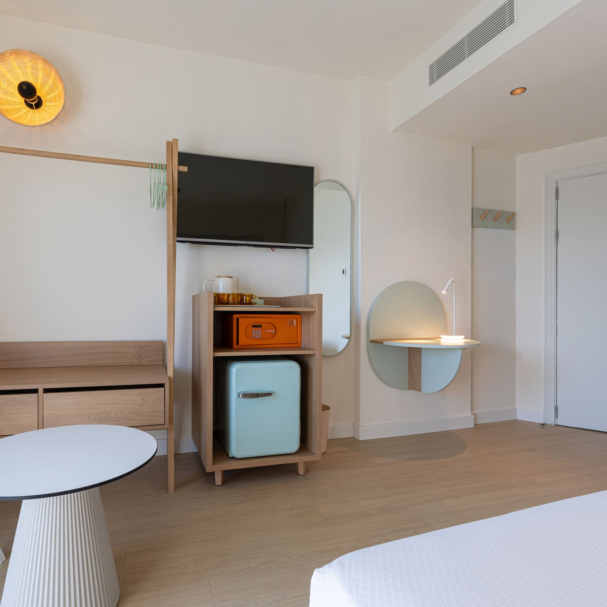 Bright, modern hotel room with minimalist light wood furniture, TV, mini-fridge.