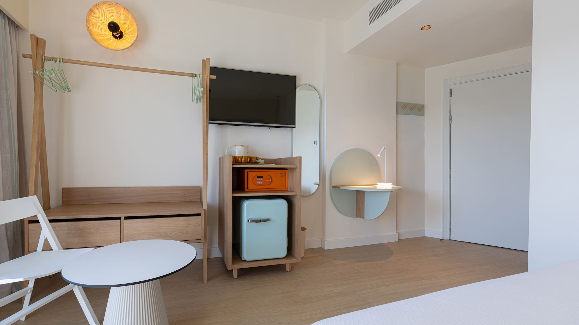 Bright, modern hotel room with minimalist light wood furniture, TV, mini-fridge.