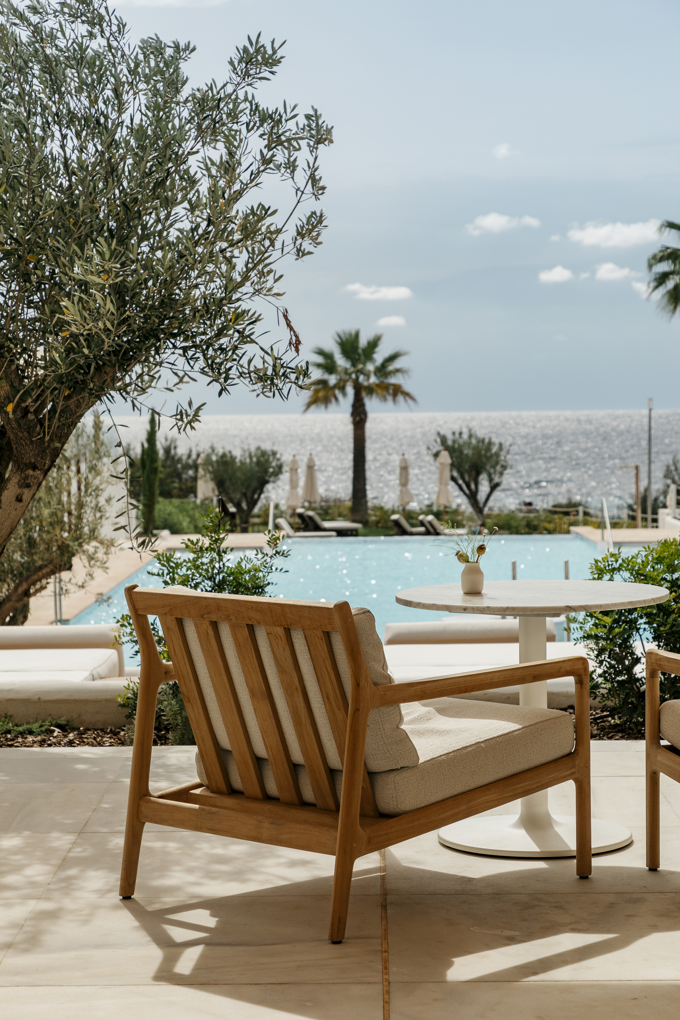 Outdoor patio with wooden armchair, marble table, pool, and sparkling sea view.