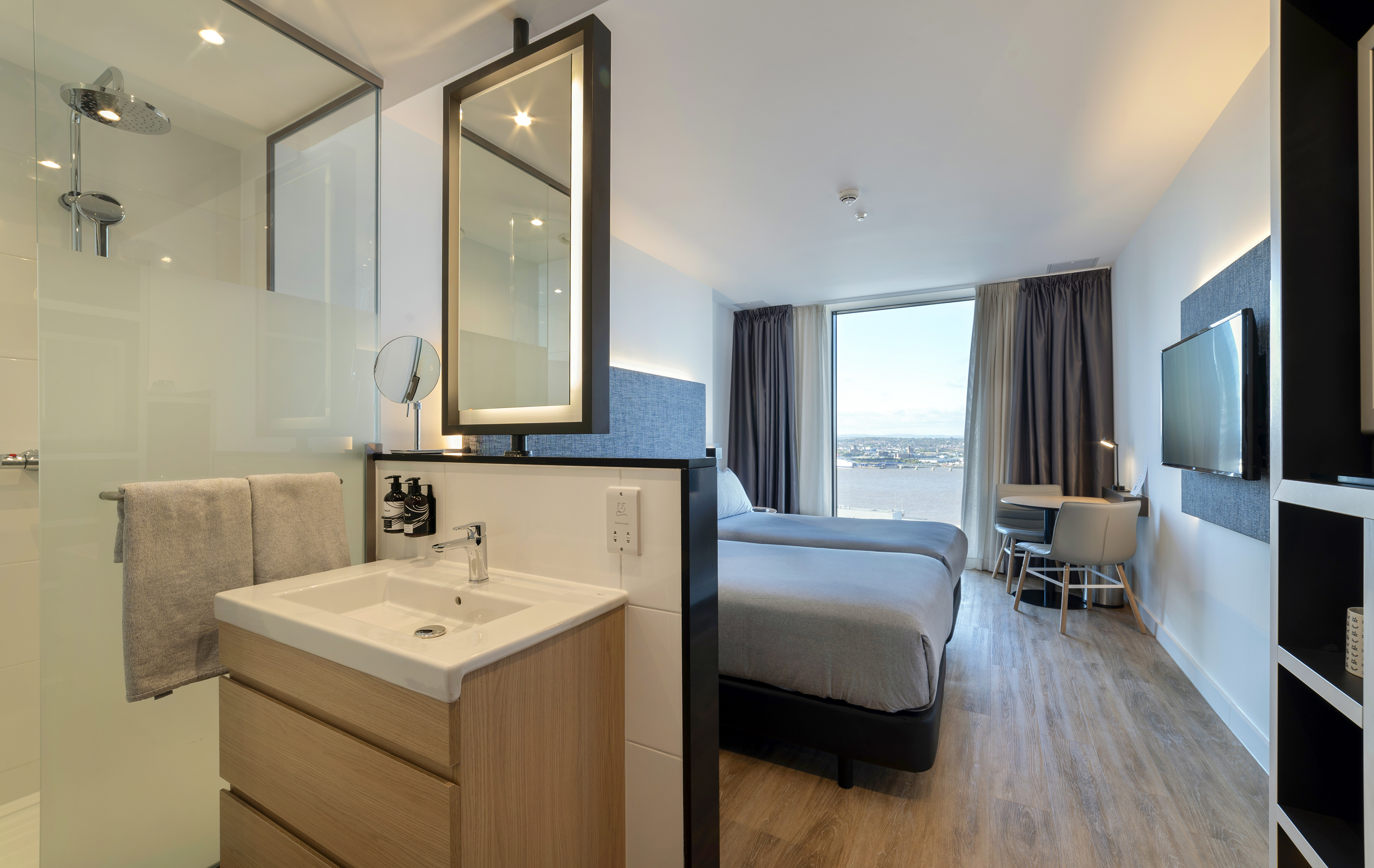 Modern hotel room with twin beds, city view, and open-plan bathroom with shower.