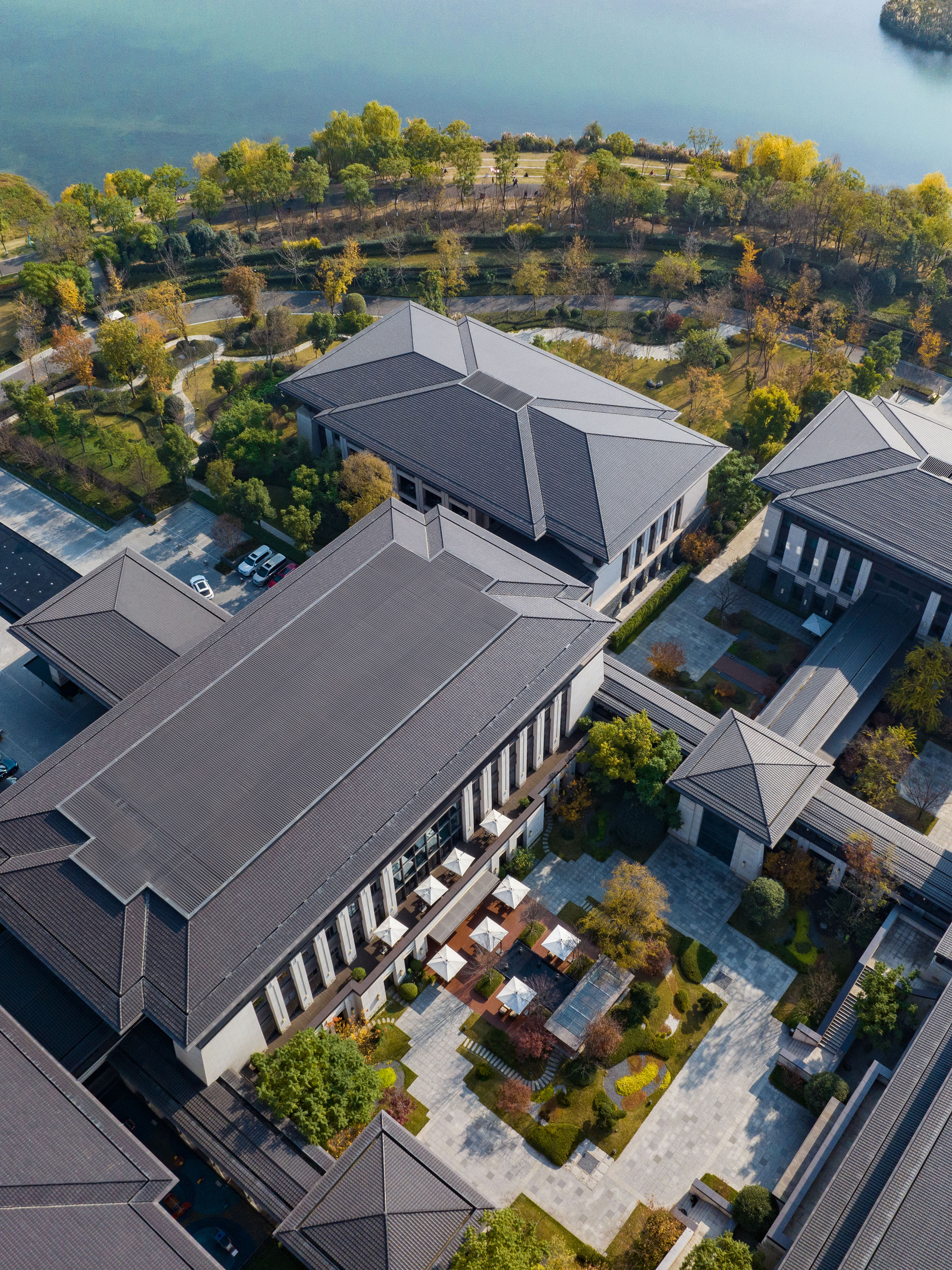 Aerial view of modern buildings with dark roofs, landscaped gardens, autumn trees by a lake.