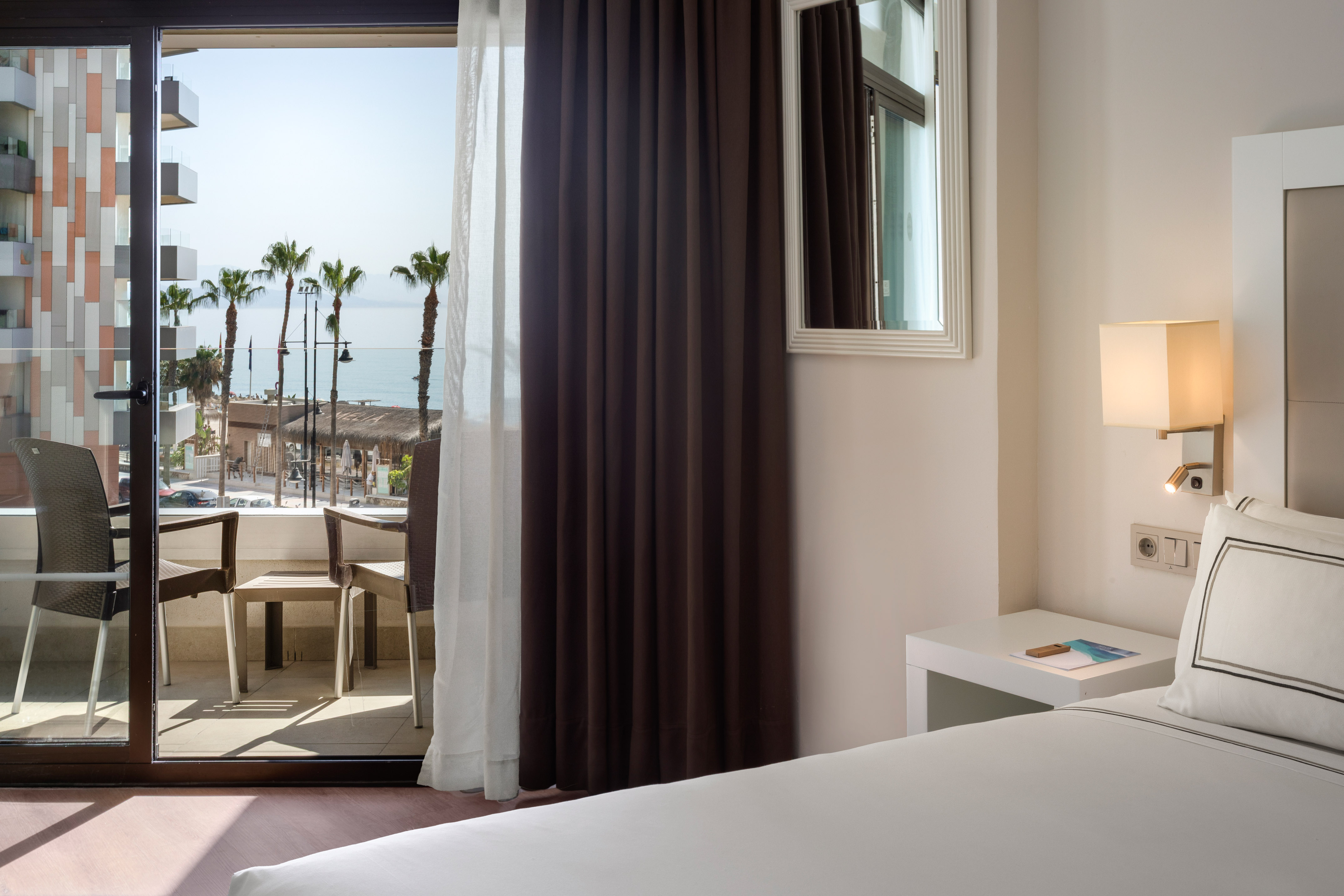Modern hotel room with balcony, sea view, palm trees, and beach.