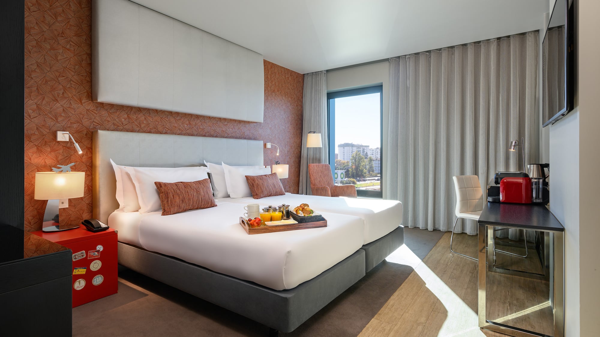 Modern hotel room with breakfast in bed, large window, city view, and desk.