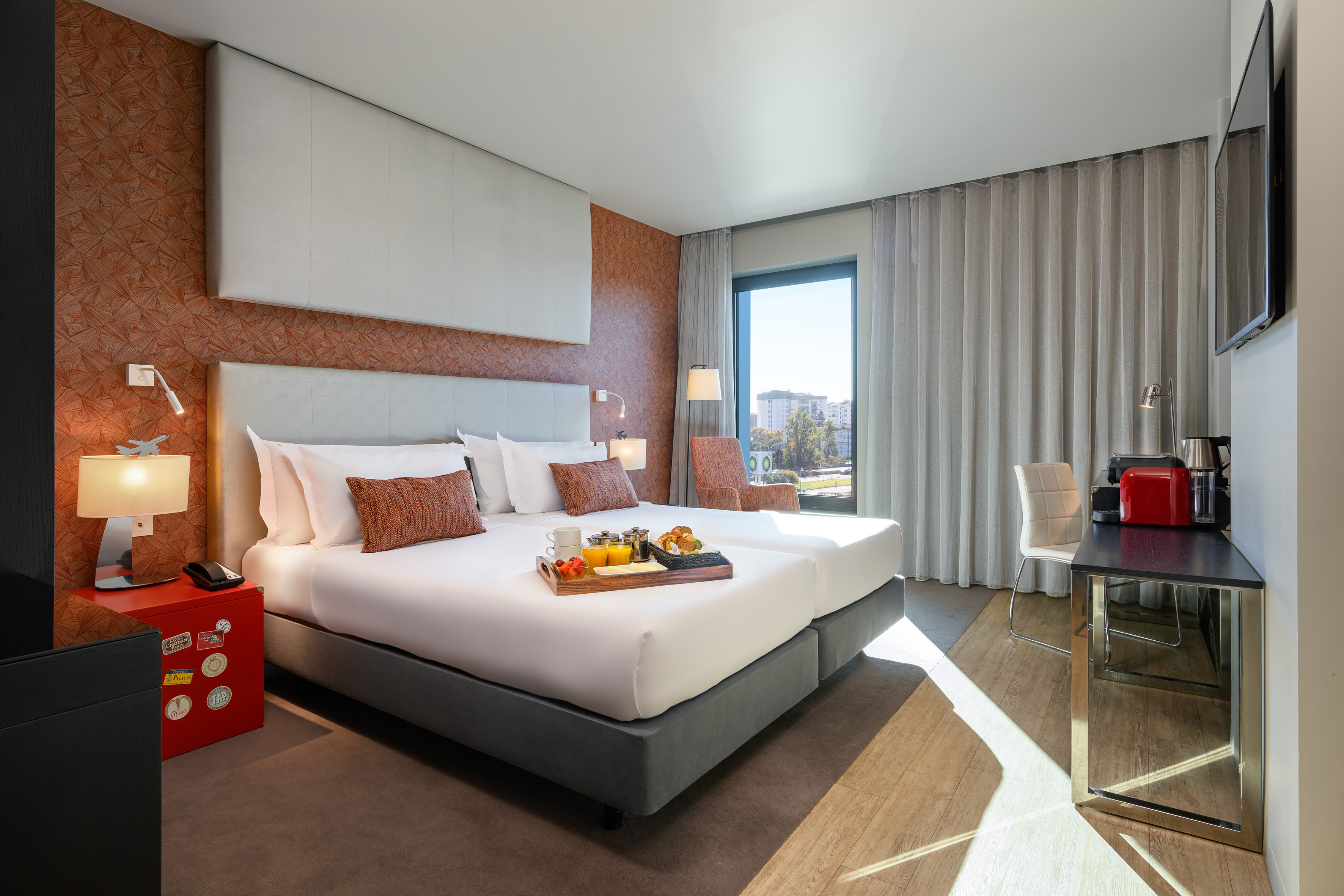 Modern hotel room with breakfast in bed, large window, city view, and desk.
