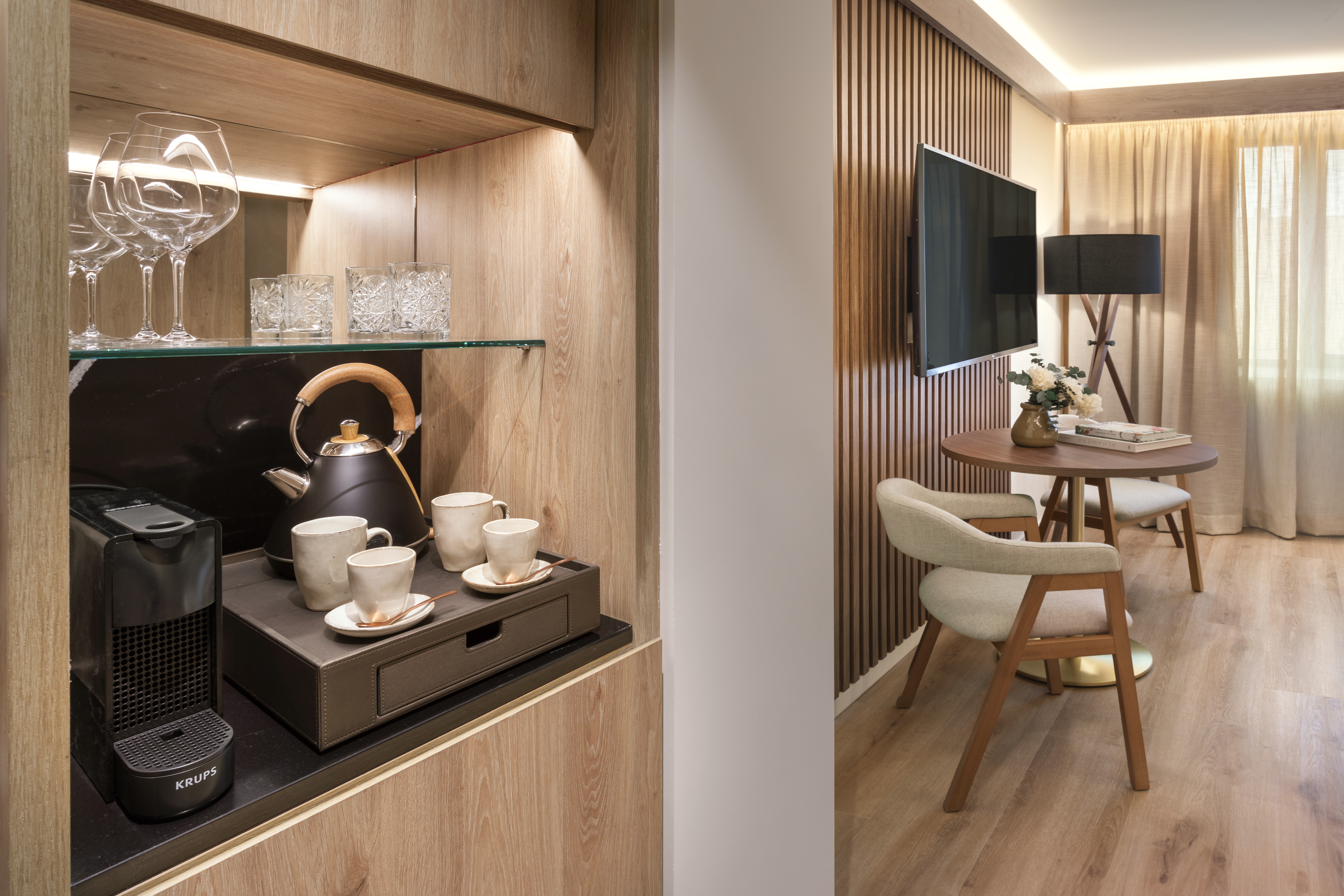 Modern hotel room: coffee station, kettle, glasses, and seating with TV.