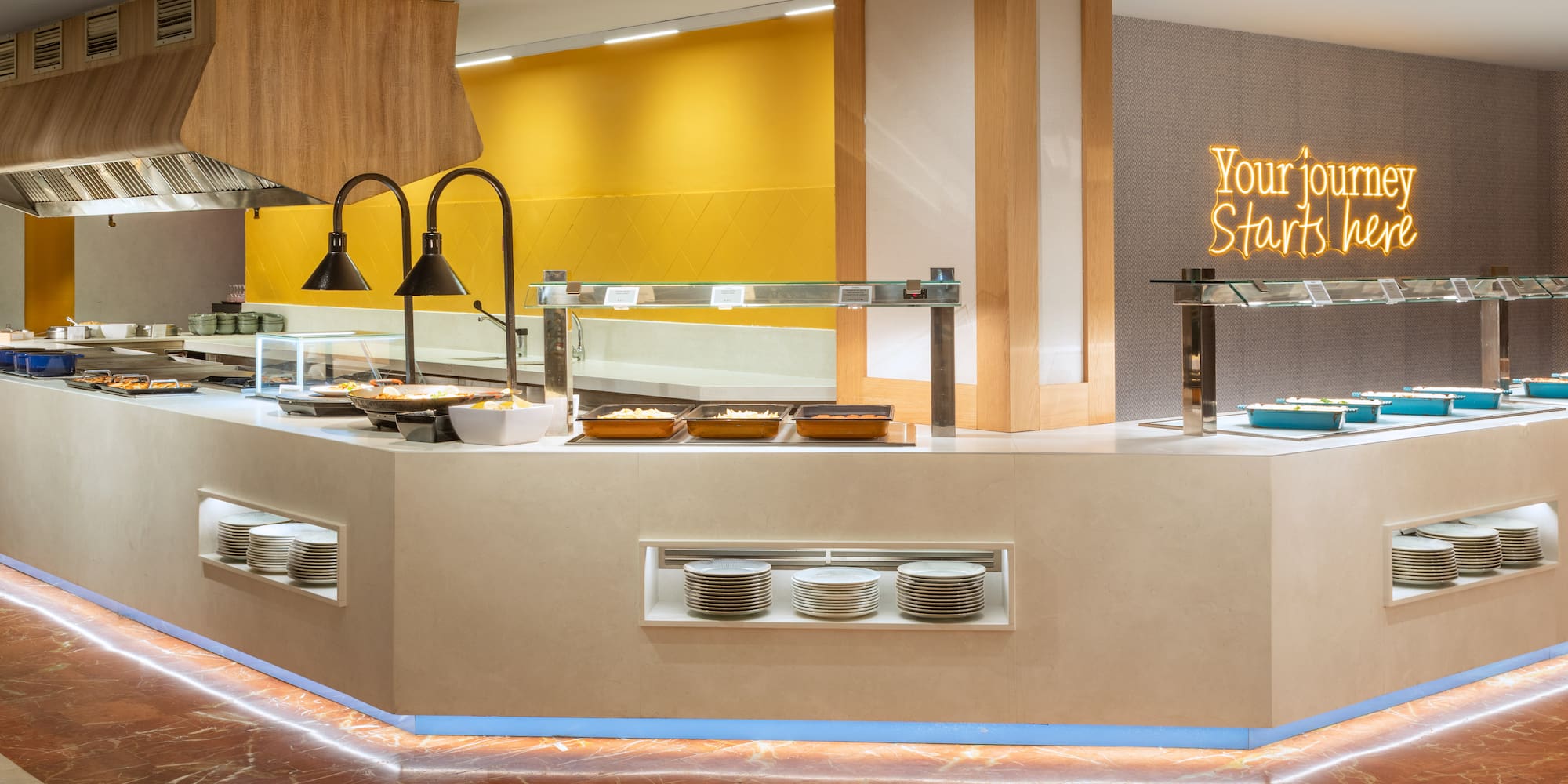 Modern hotel buffet line with hot dishes, stacked plates, and "Your journey Starts here" neon sign.