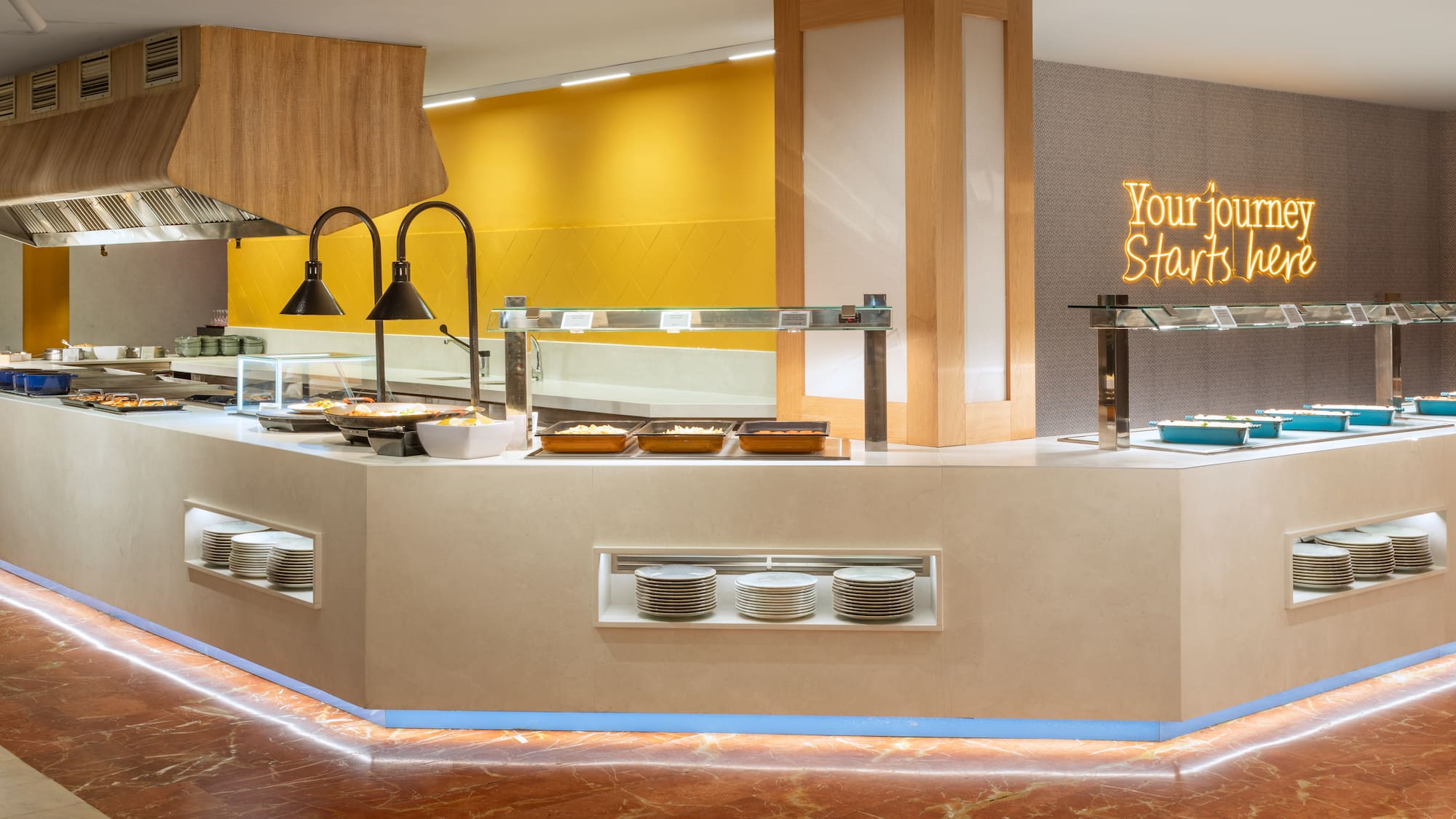 Modern hotel buffet line with hot dishes, stacked plates, and "Your journey Starts here" neon sign.
