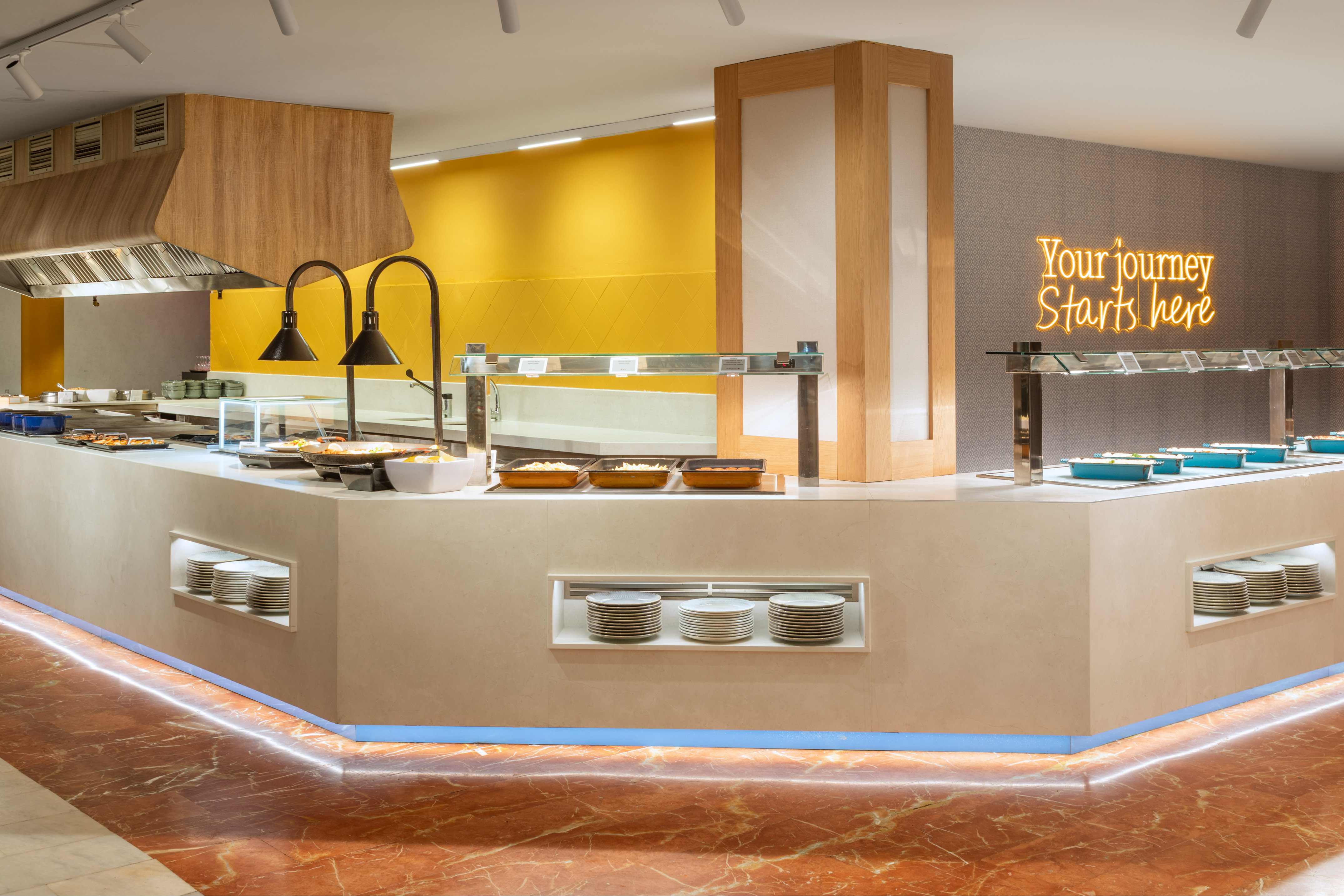 Modern hotel buffet line with hot dishes, stacked plates, and "Your journey Starts here" neon sign.