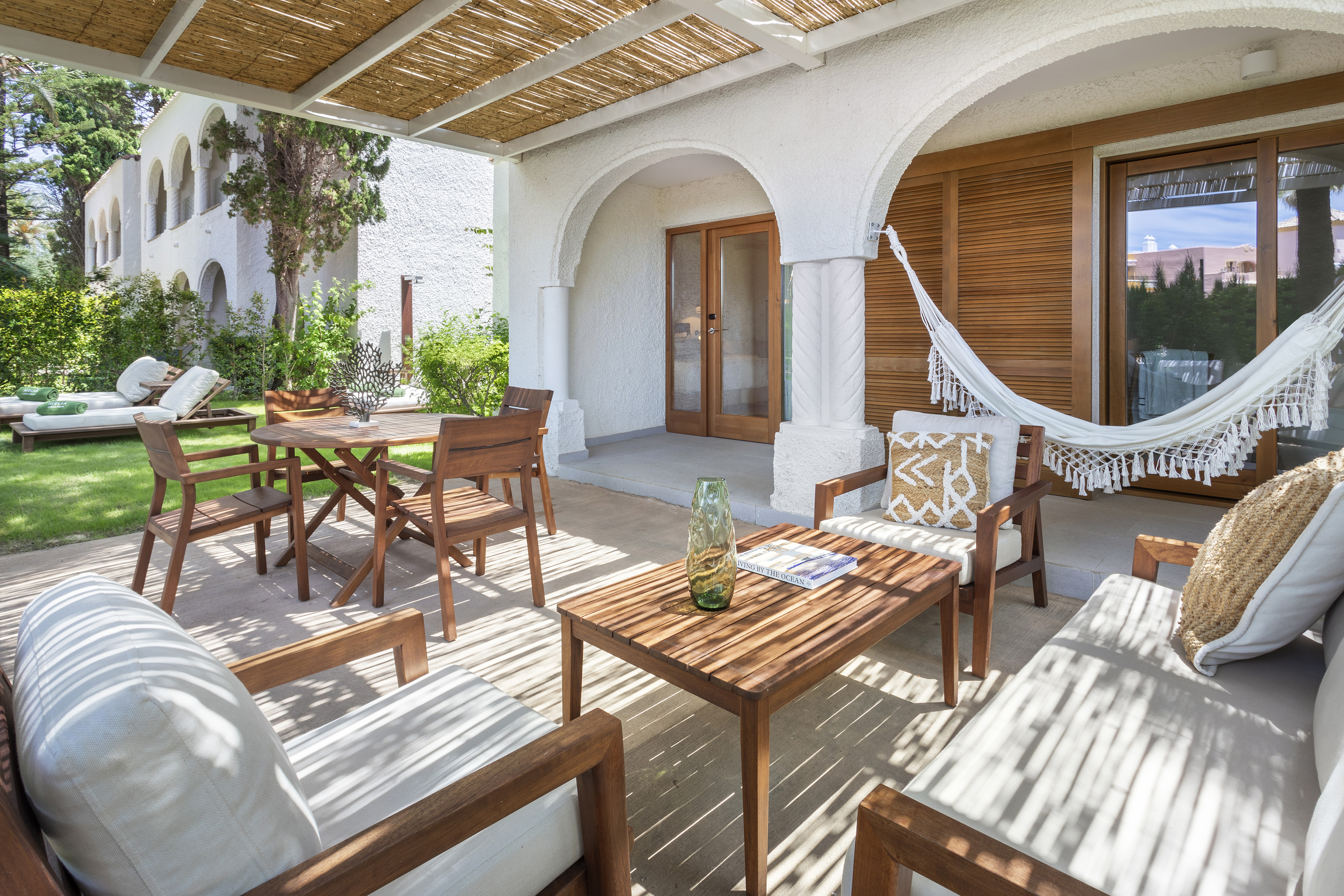 Resort patio with wooden outdoor furniture, white hammock, and Mediterranean arches.