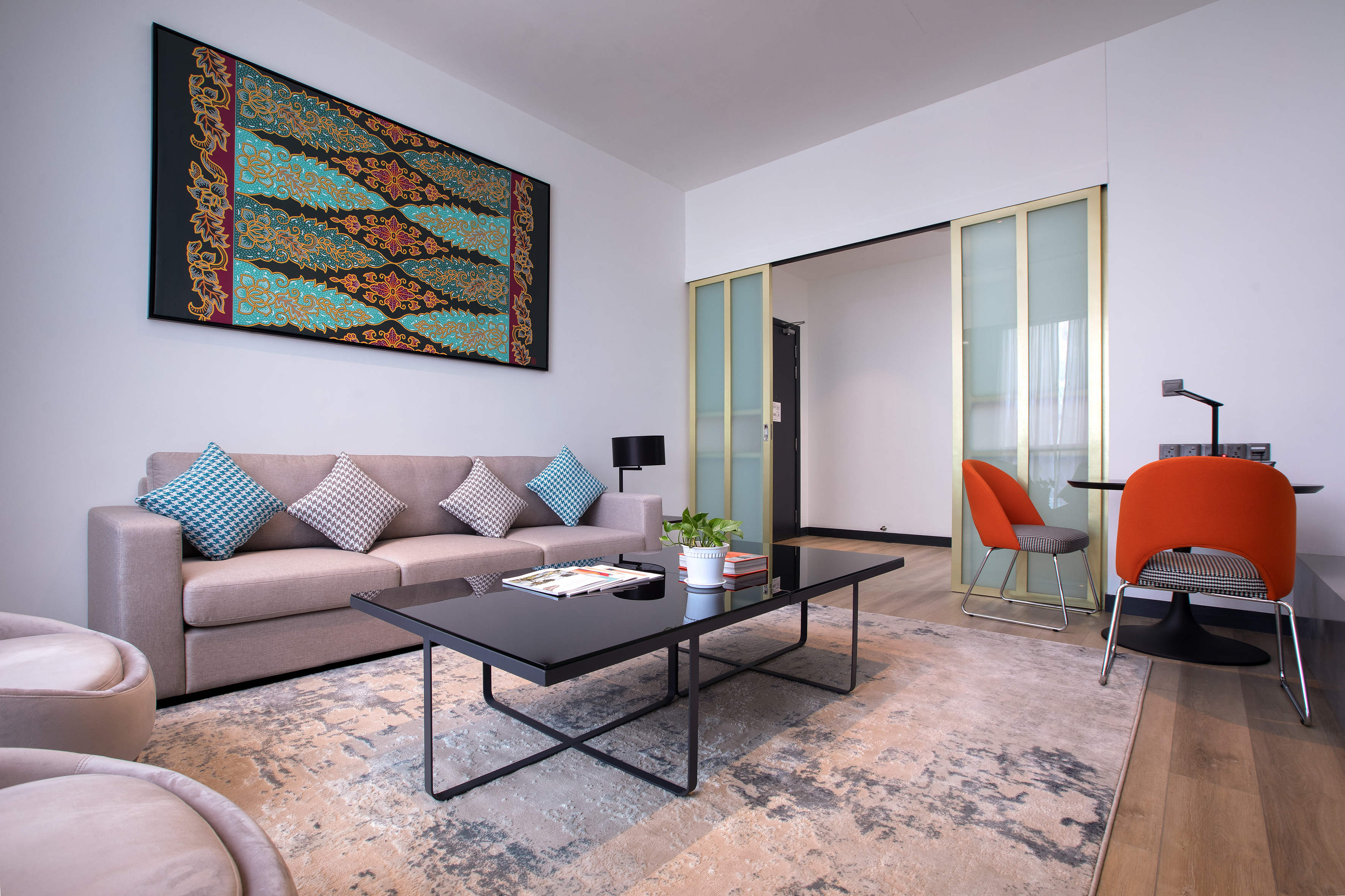 Modern living room with sofa, colorful batik wall art, black coffee table, orange chairs.