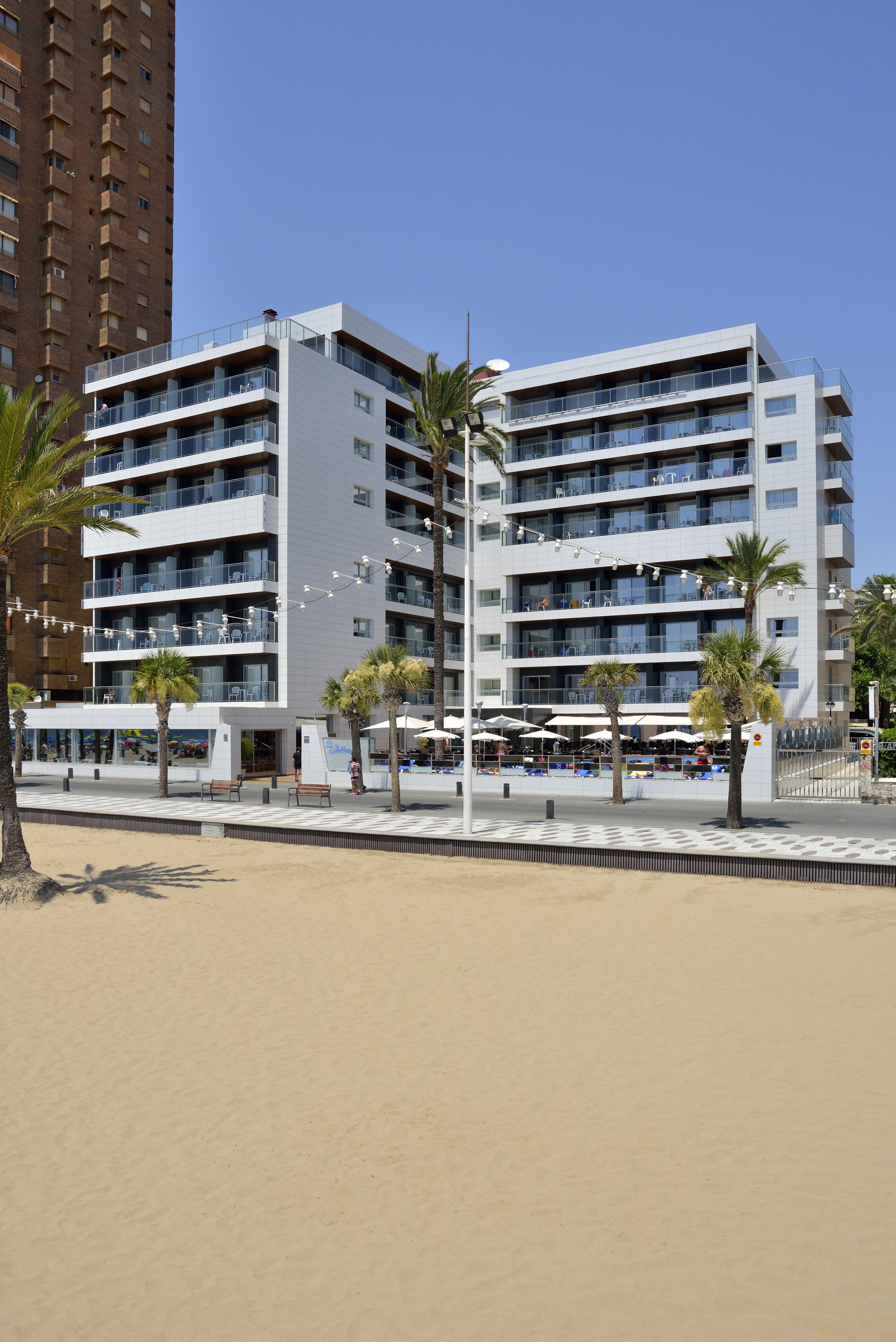 Modern beachfront hotel with glass balconies, palm trees, and sandy beach.