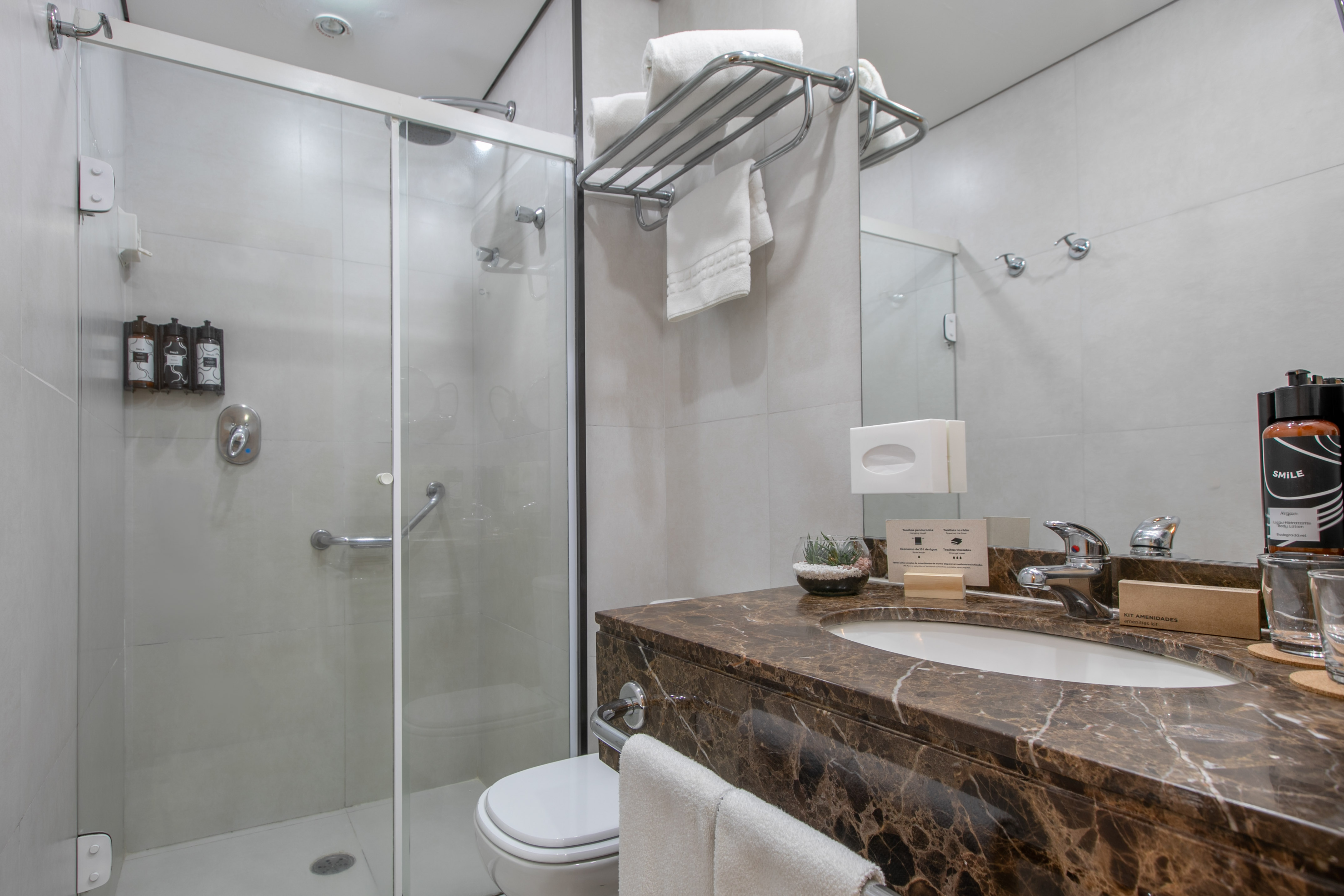 Modern hotel bathroom with glass shower, brown marble vanity, toilet, and amenities.